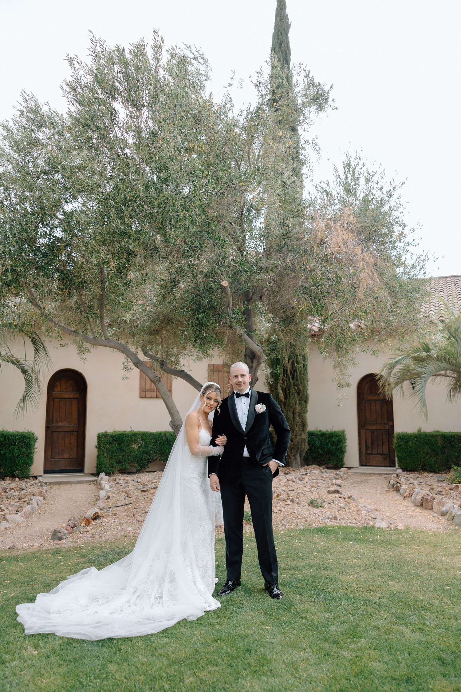 Bride and groom pose together on green lawn before rustic venue with arched doors and olive tree.
