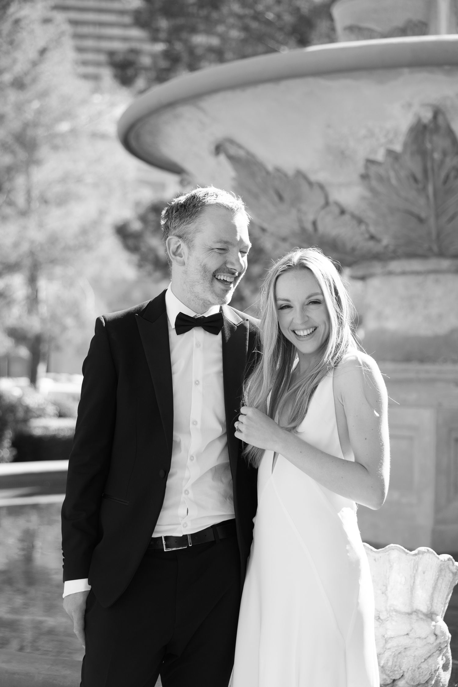 Black and white photo of a smiling couple in formal wedding attire posing in front of an ornate stone fountain.