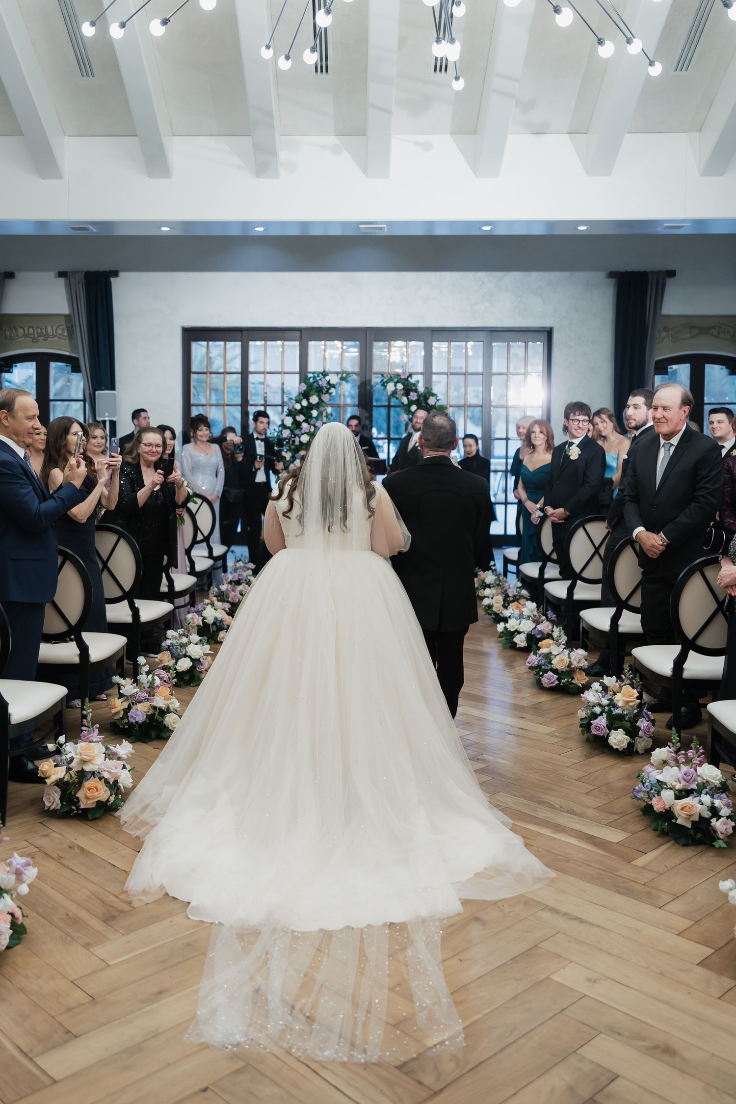 Bride in a white ball gown and veil walks down the aisle at an elegant indoor wedding ceremony with floral arrangements.