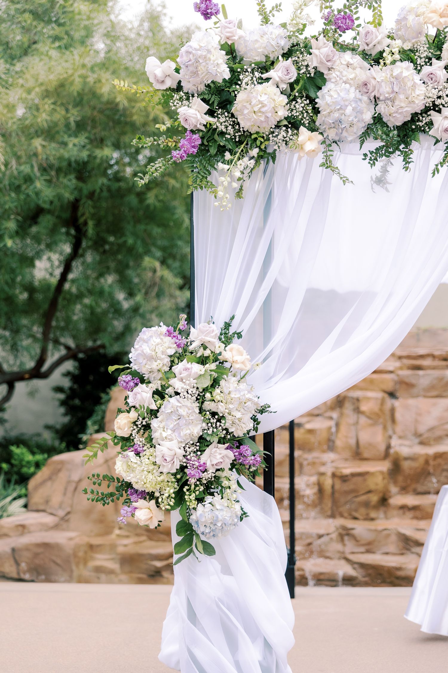 Elegant wedding arch with white draping and lush floral arrangements featuring white hydrangeas and purple accents outdoors.