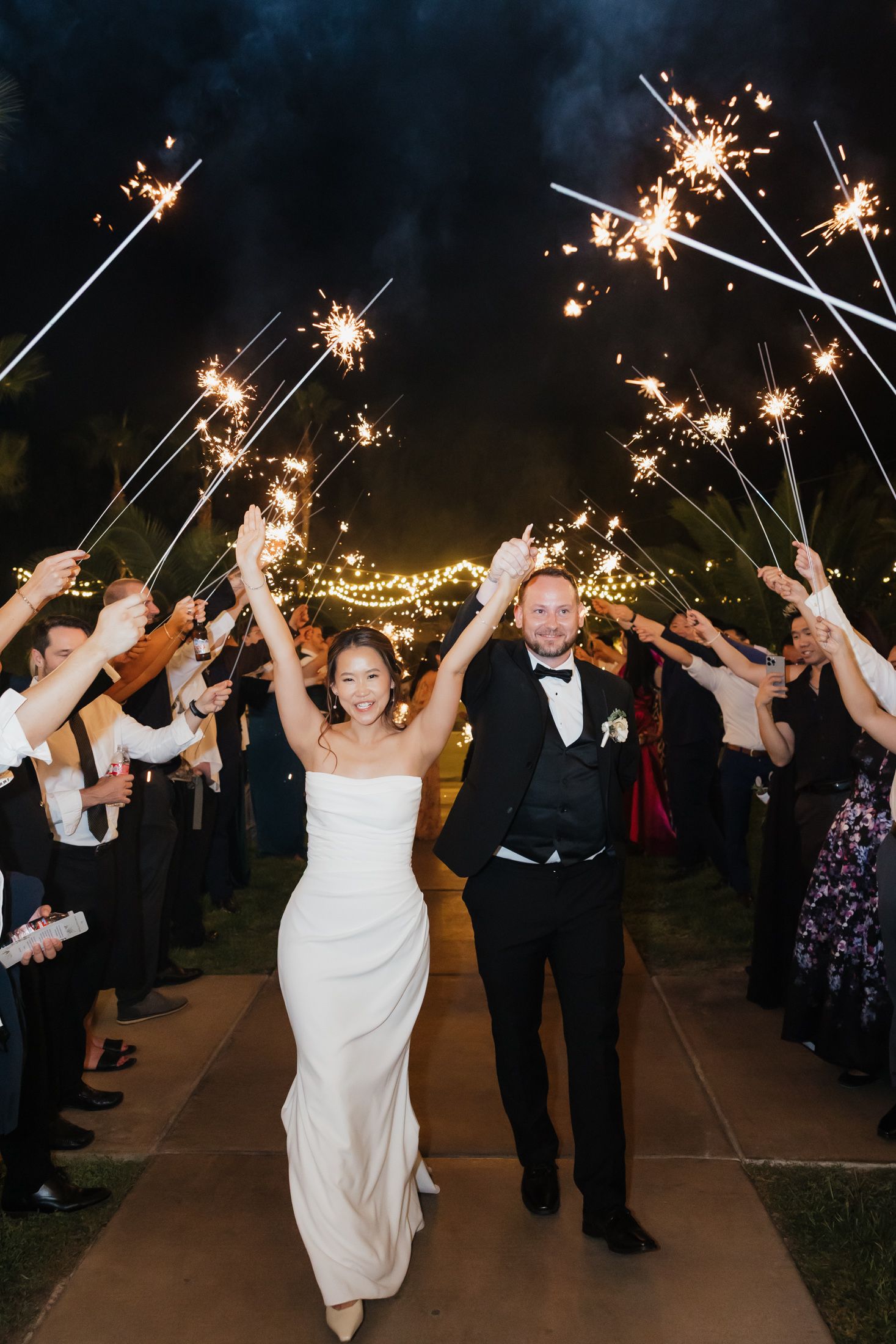 Wedding couple makes grand sparkler exit through tunnel of guests holding lit sparklers at nighttime celebration.