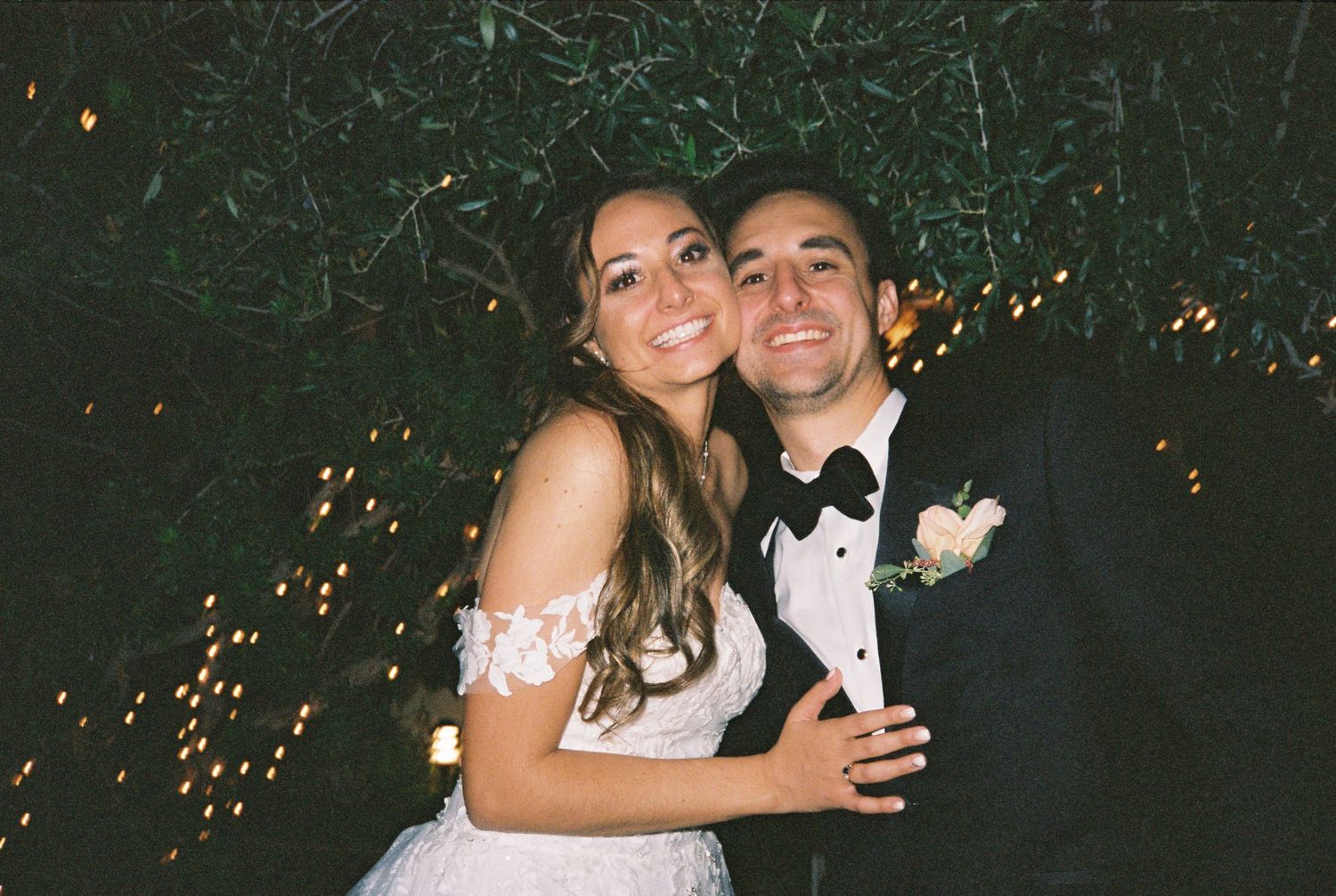 Happy bride and groom embracing outdoors at night with twinkling lights behind them at their wedding reception.