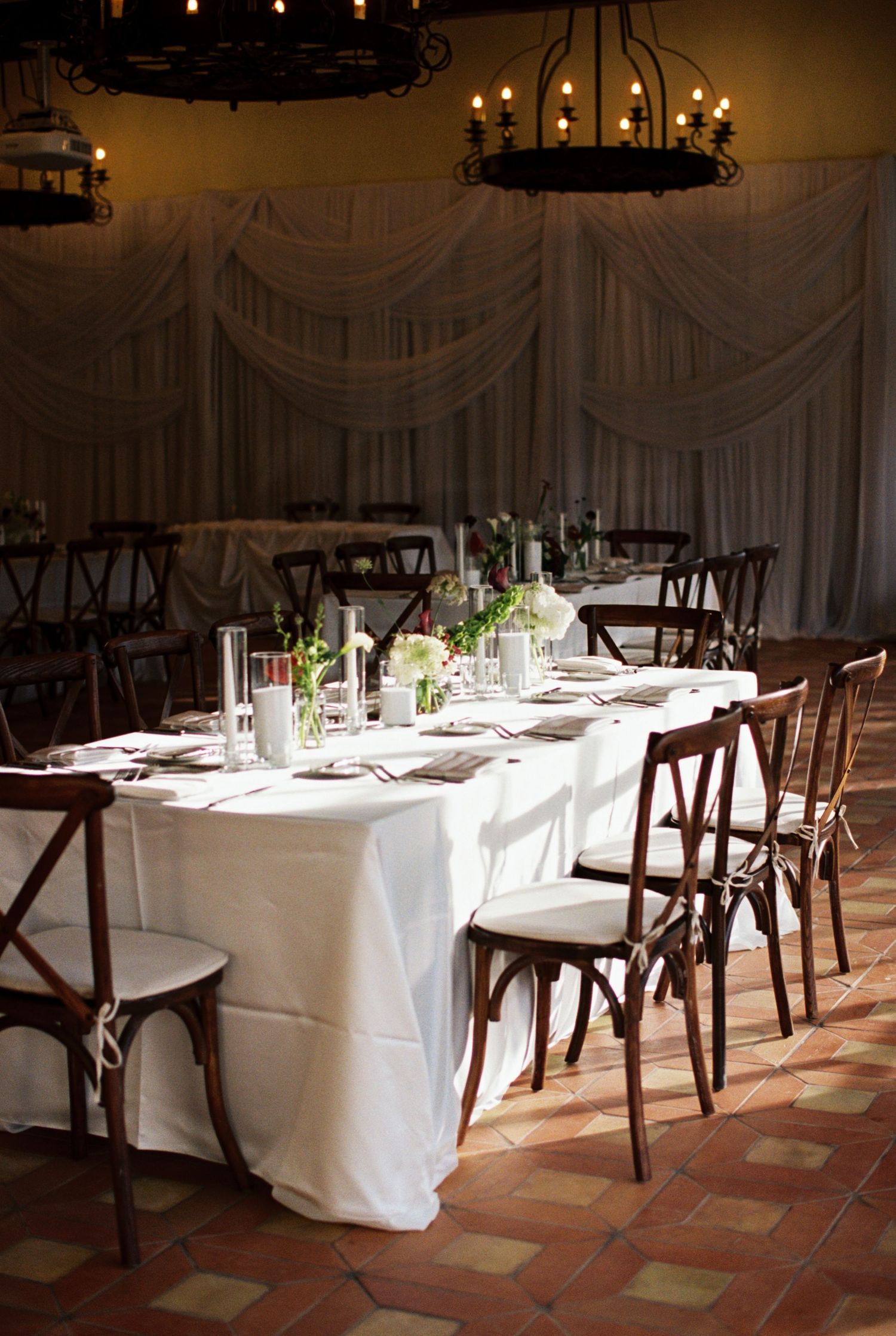 Elegant wedding reception hall with white draped tables, rustic cross-back chairs, floral centerpieces, and wrought iron chandeliers.
