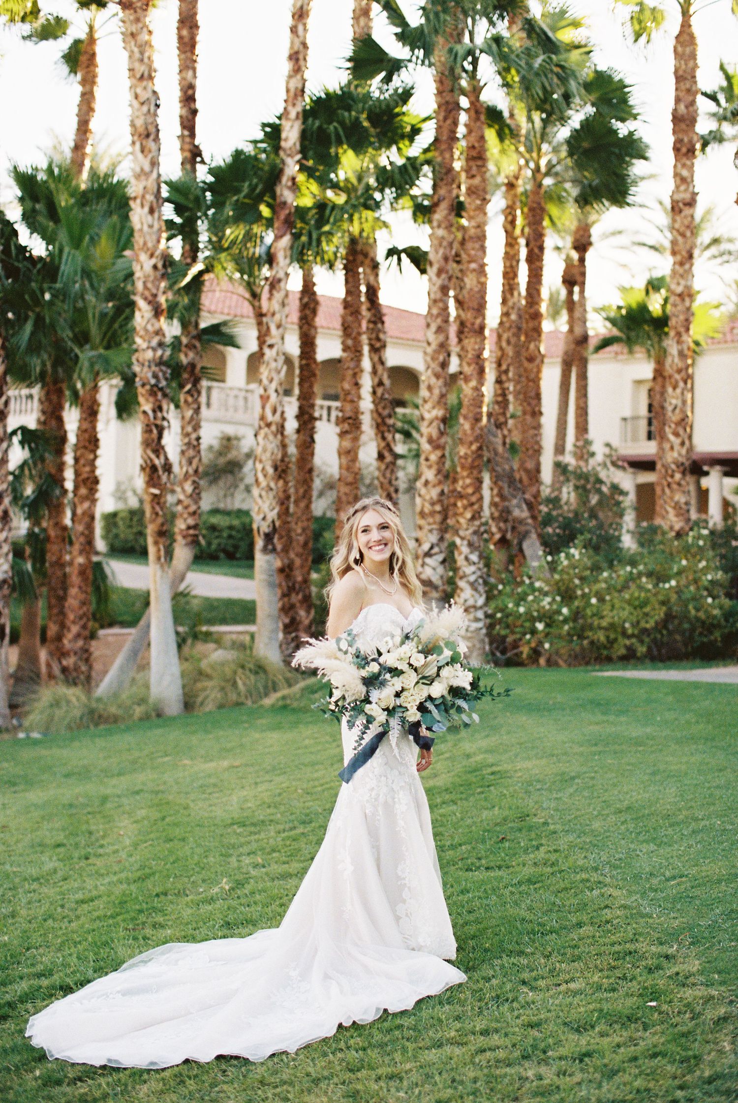 Smiling bride in white lace gown with long train holds bouquet on green lawn surrounded by tall palm trees.