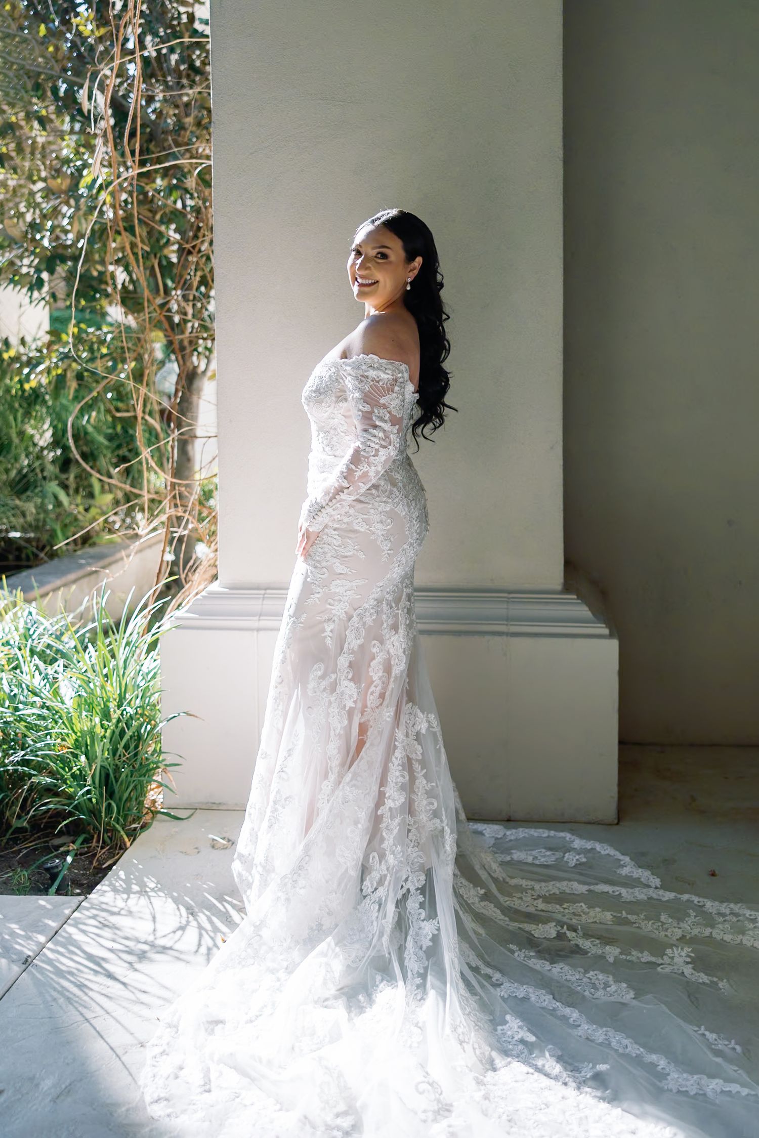 Bride in off-shoulder lace wedding gown with long train poses by elegant column in sunlit outdoor setting.