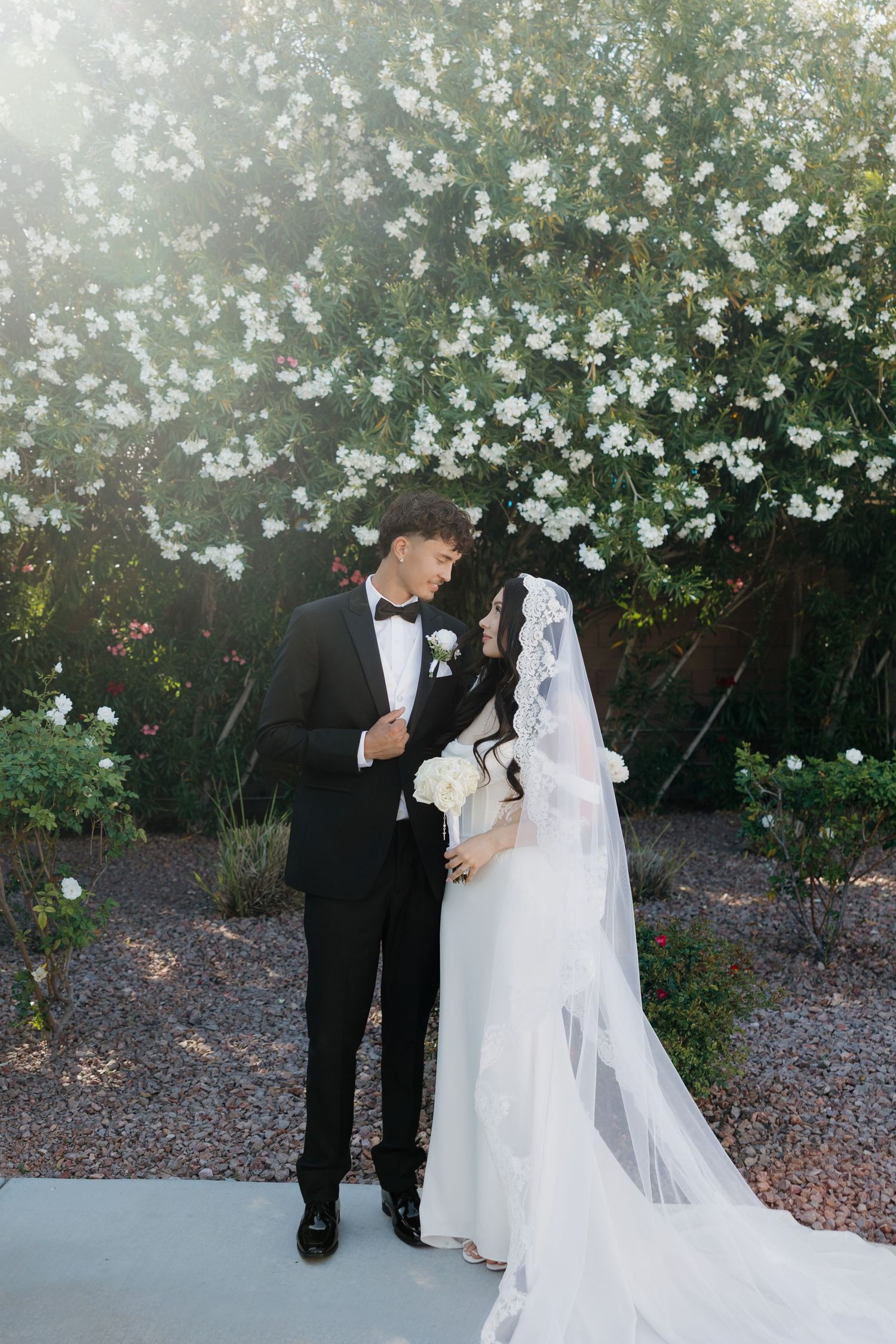 Bride and groom share a romantic moment in a garden, with white blooming flowers backdrop, bride in veil holding bouquet.