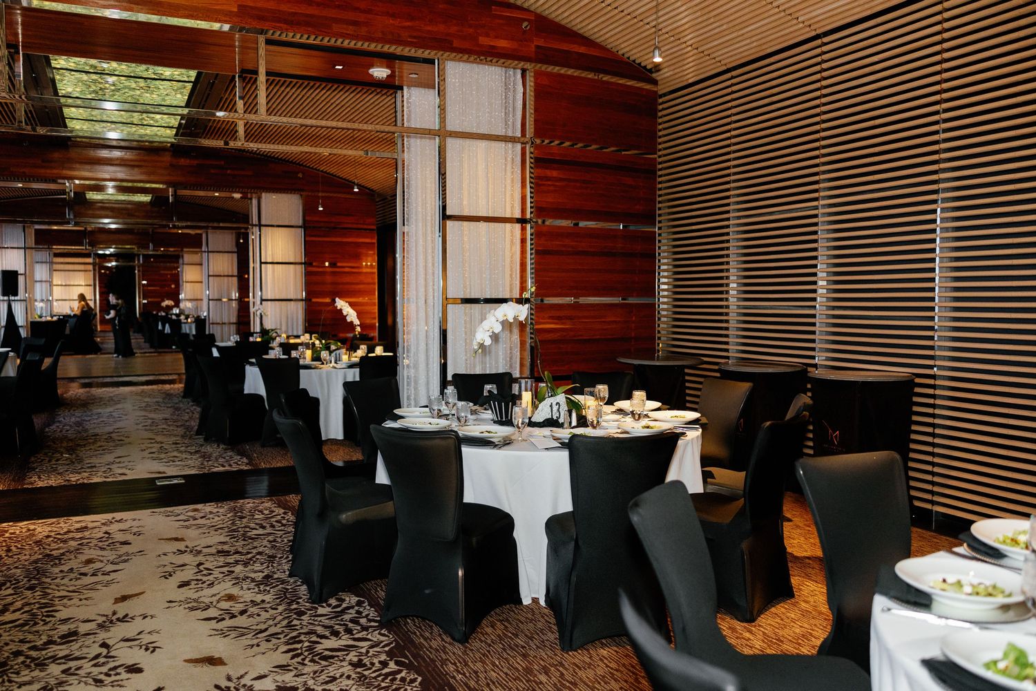 Elegant restaurant dining room with black chairs, white tablecloths, floral centerpieces, and warm wood-paneled walls.