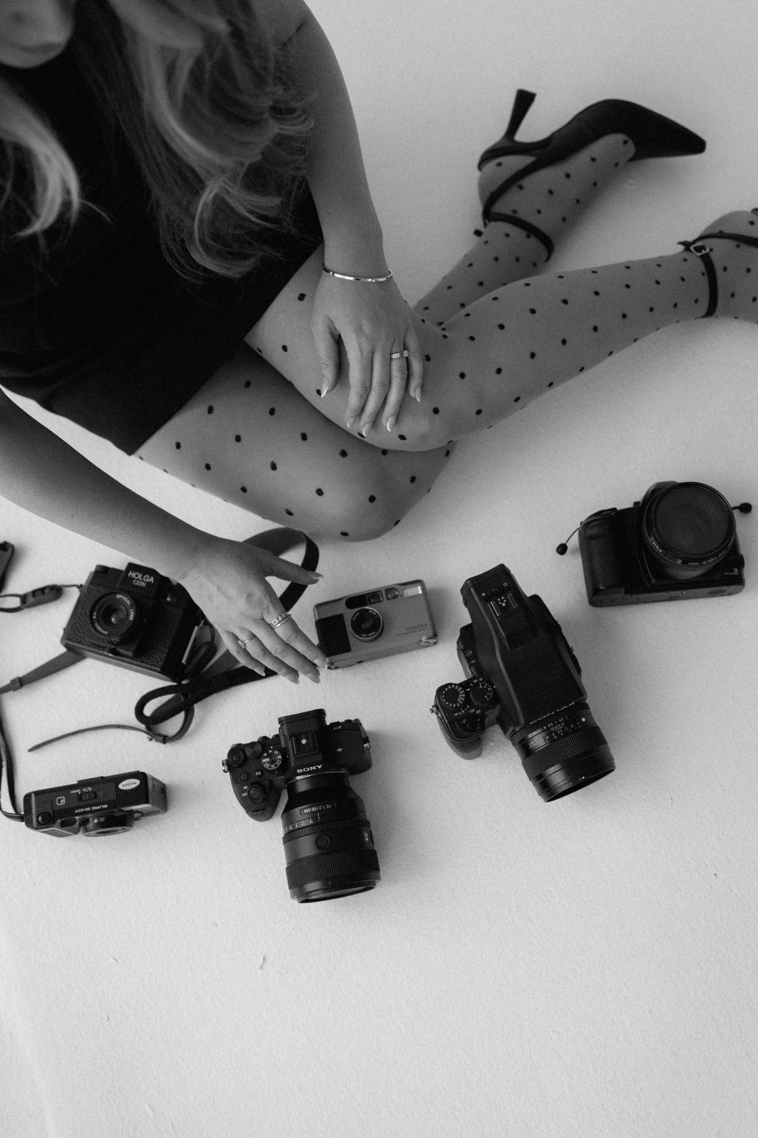 Black and white photo of woman in polka dot tights and heels sitting beside collection of vintage film cameras.