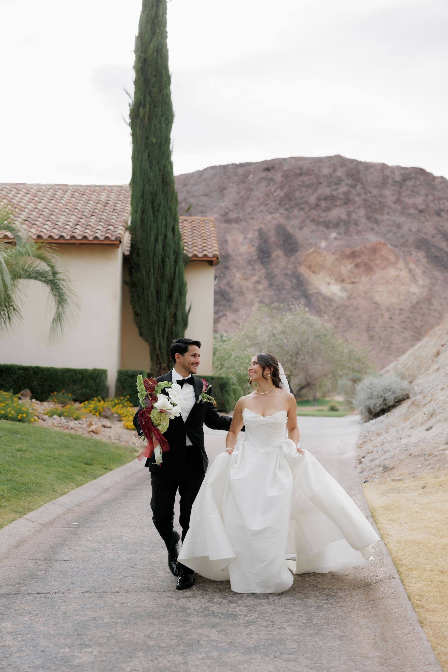 Bride in a white strapless gown and groom in a black tuxedo with a bouquet walk hand in hand along a desert path near red mountains.