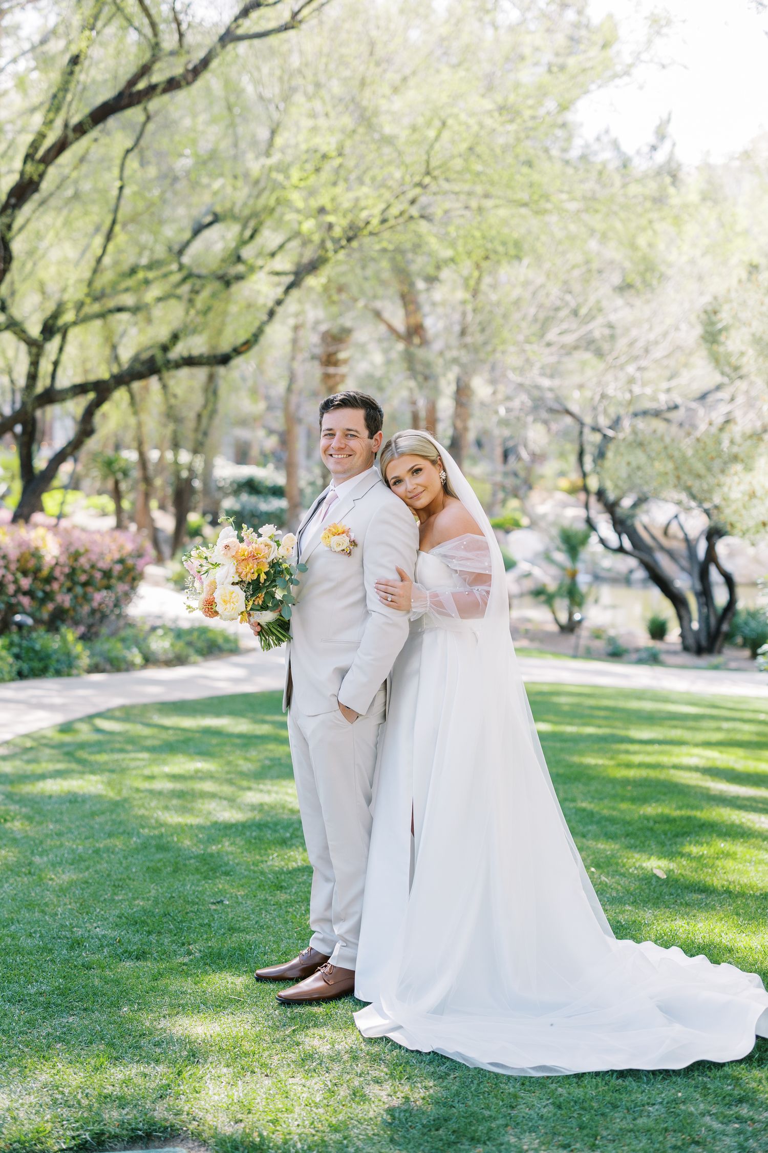 Bride in white off-shoulder gown embraces groom in tan suit holding yellow bouquet on green lawn with trees.