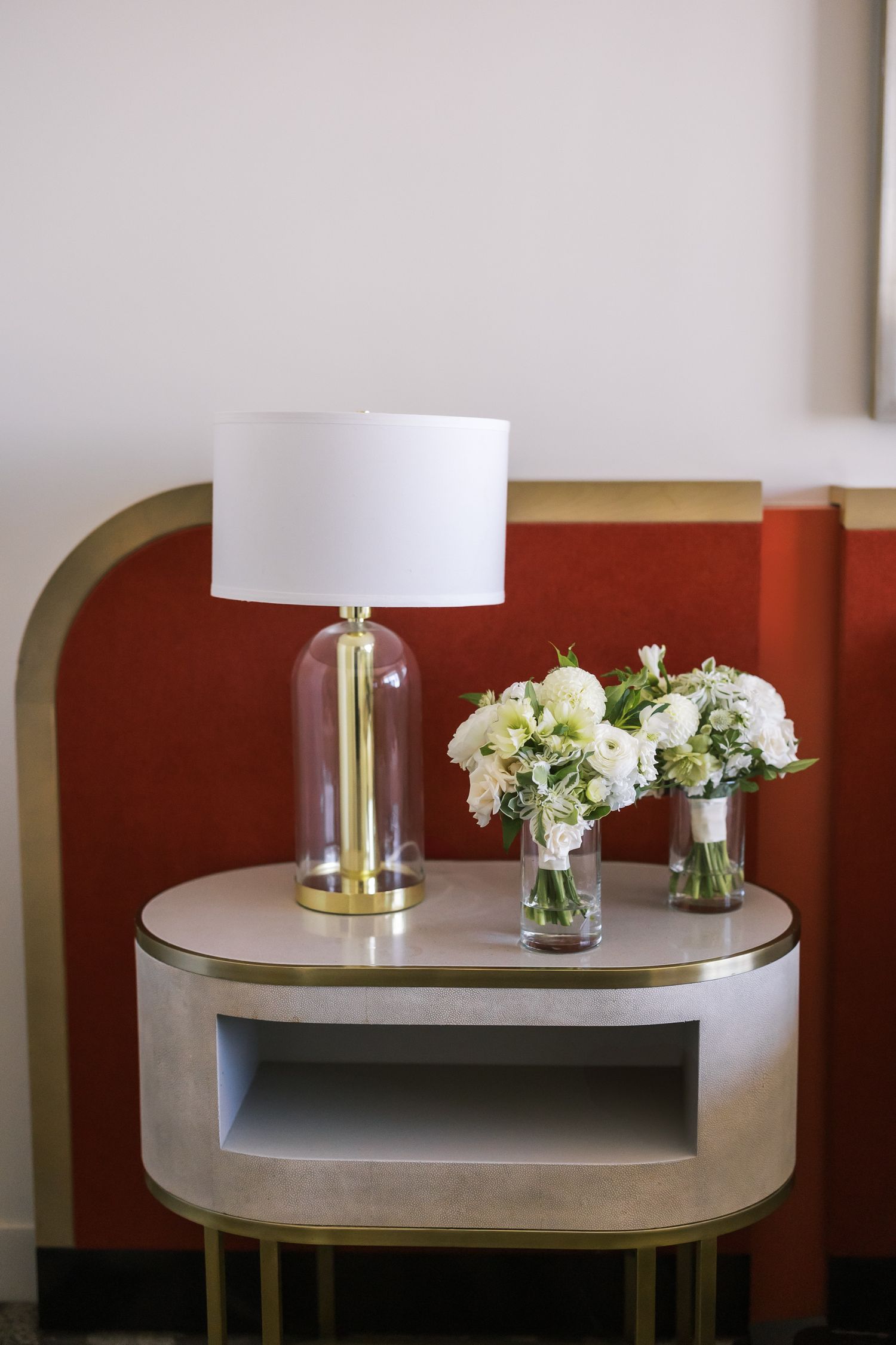 Elegant bedside table with glass and gold lamp beside white floral arrangements against a red velvet headboard.
