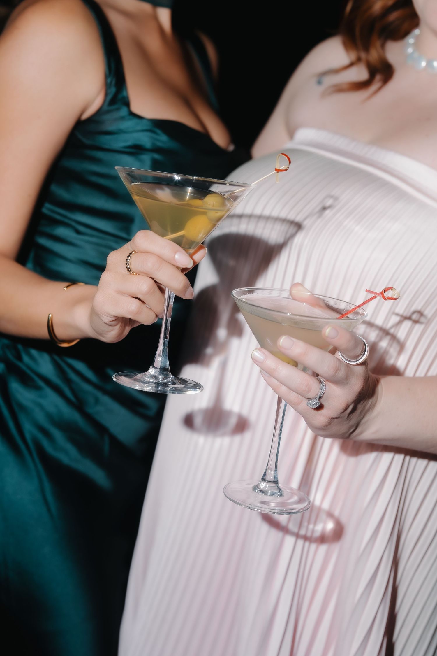 Two elegant women in formal gowns holding martini cocktail glasses at a party event.