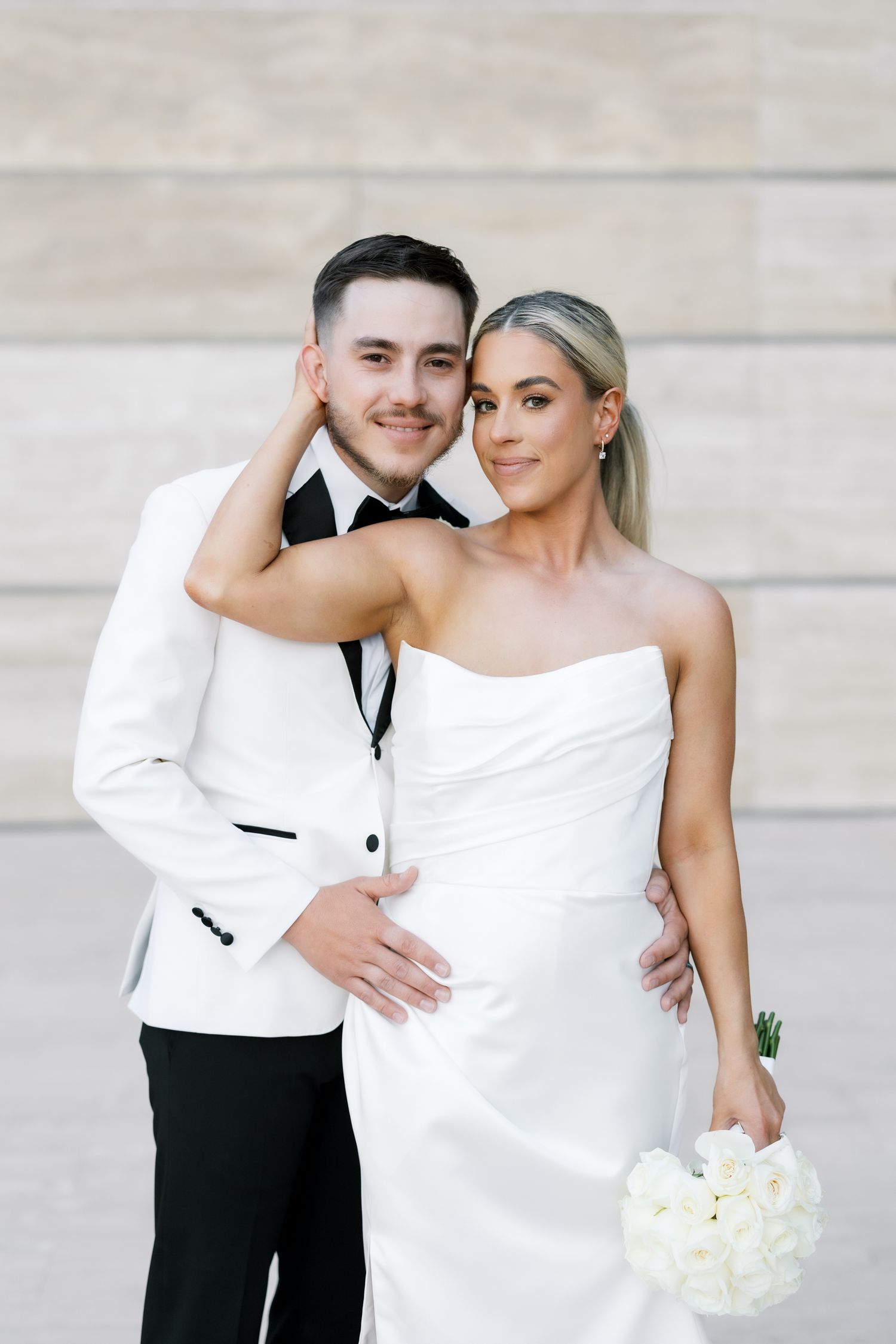 Bride and groom posing closely together, bride holding white bouquet, both in elegant white attire.