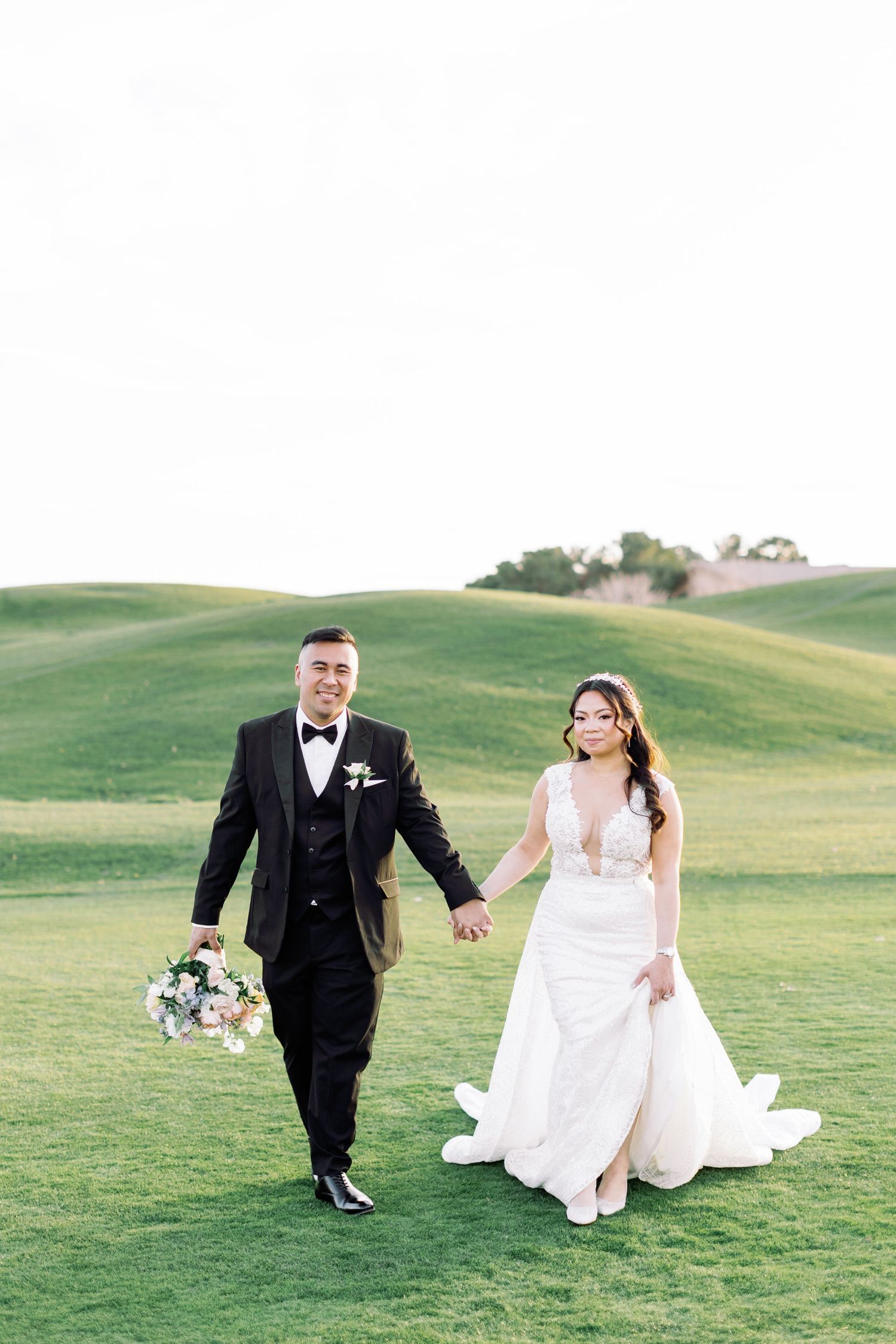Bride and groom walk hand-in-hand on lush green golf course, she in white gown, he in black tuxedo holding bouquet.