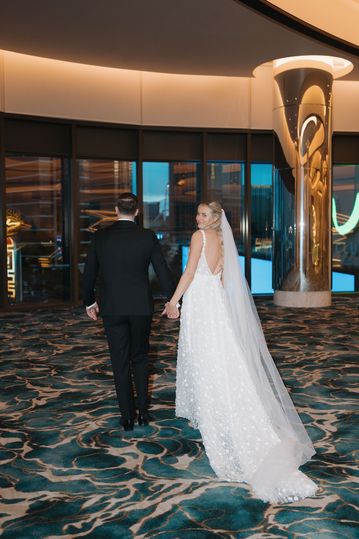 Bride and groom walk hand in hand through luxurious hotel lobby, bride wearing stunning backless wedding gown with veil.