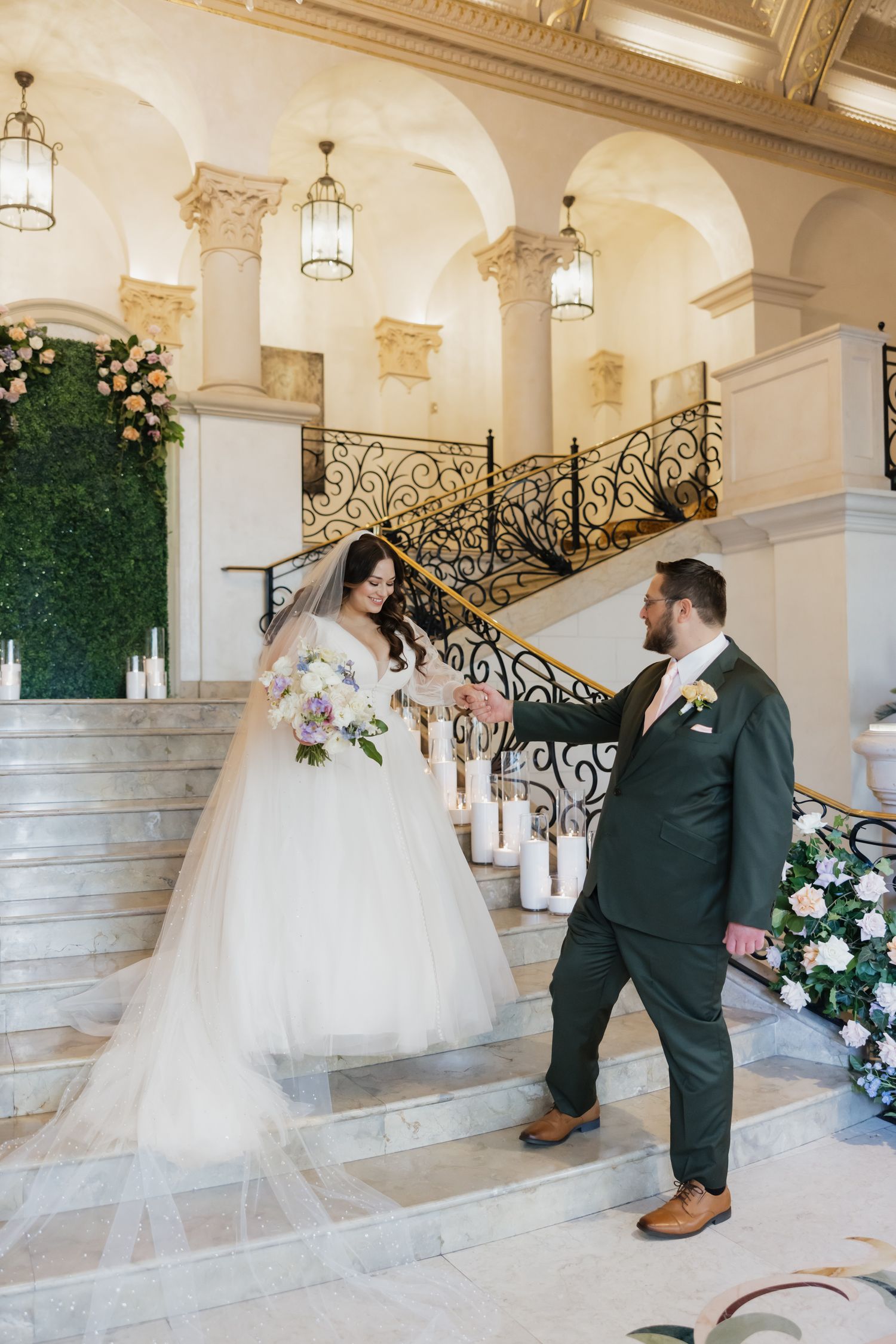 Bride and groom holding hands on grand staircase in elegant venue, bride in tulle gown with bouquet.