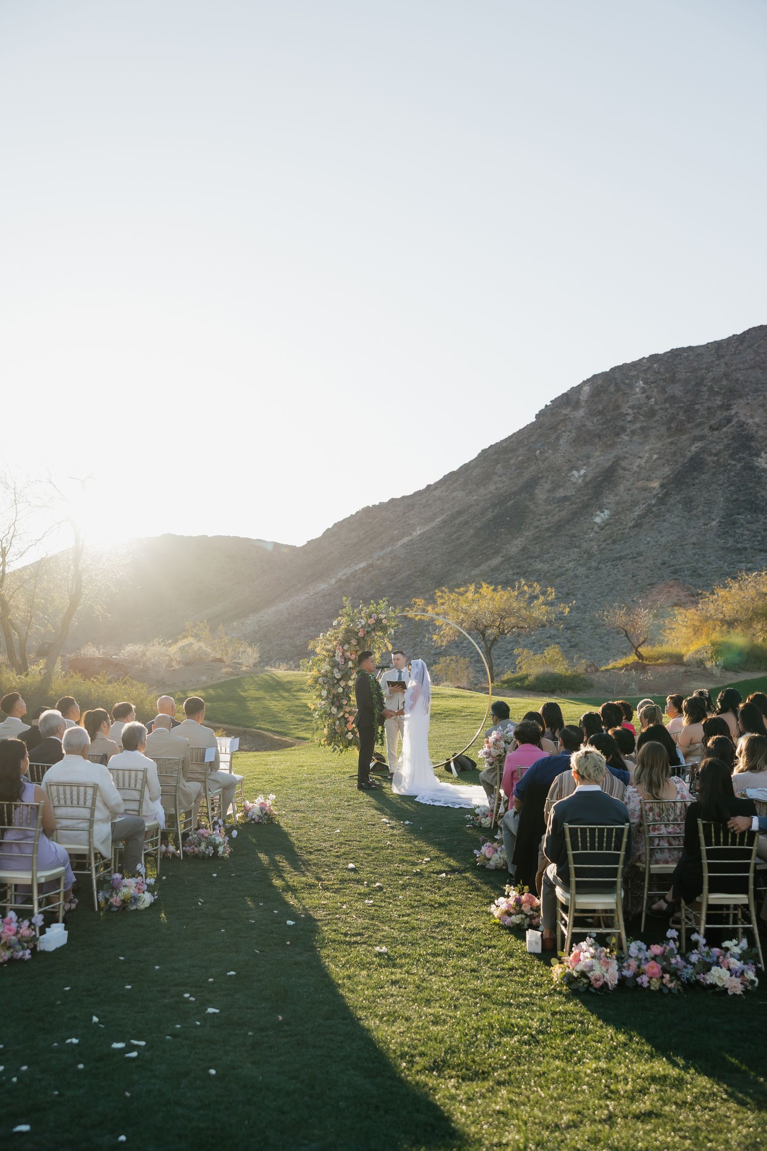 Outdoor wedding ceremony on green lawn with mountain backdrop at golden hour, guests seated in gold chairs lining floral aisle.