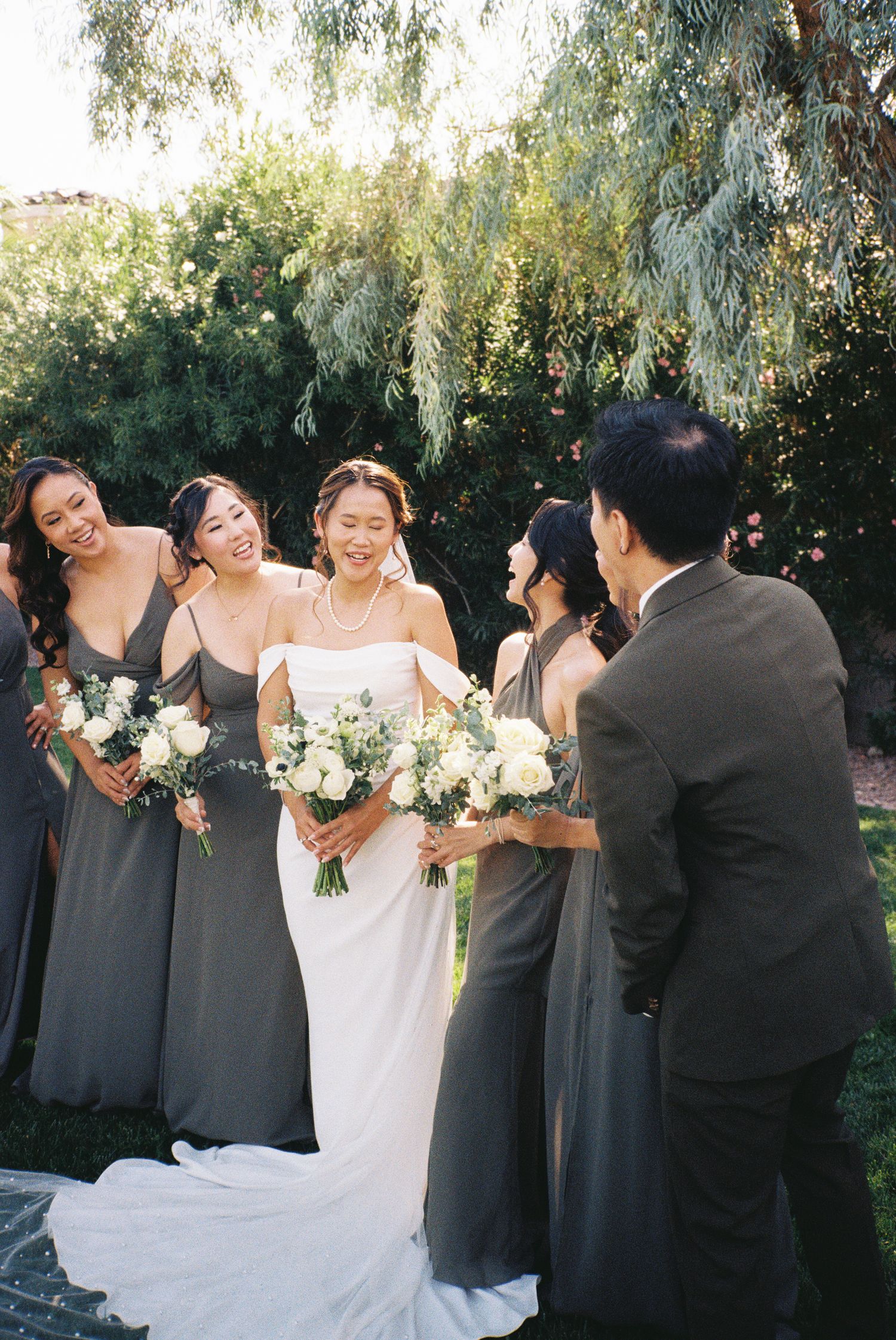 Bride in white gown holds floral bouquet surrounded by bridesmaids in gray dresses outdoors.