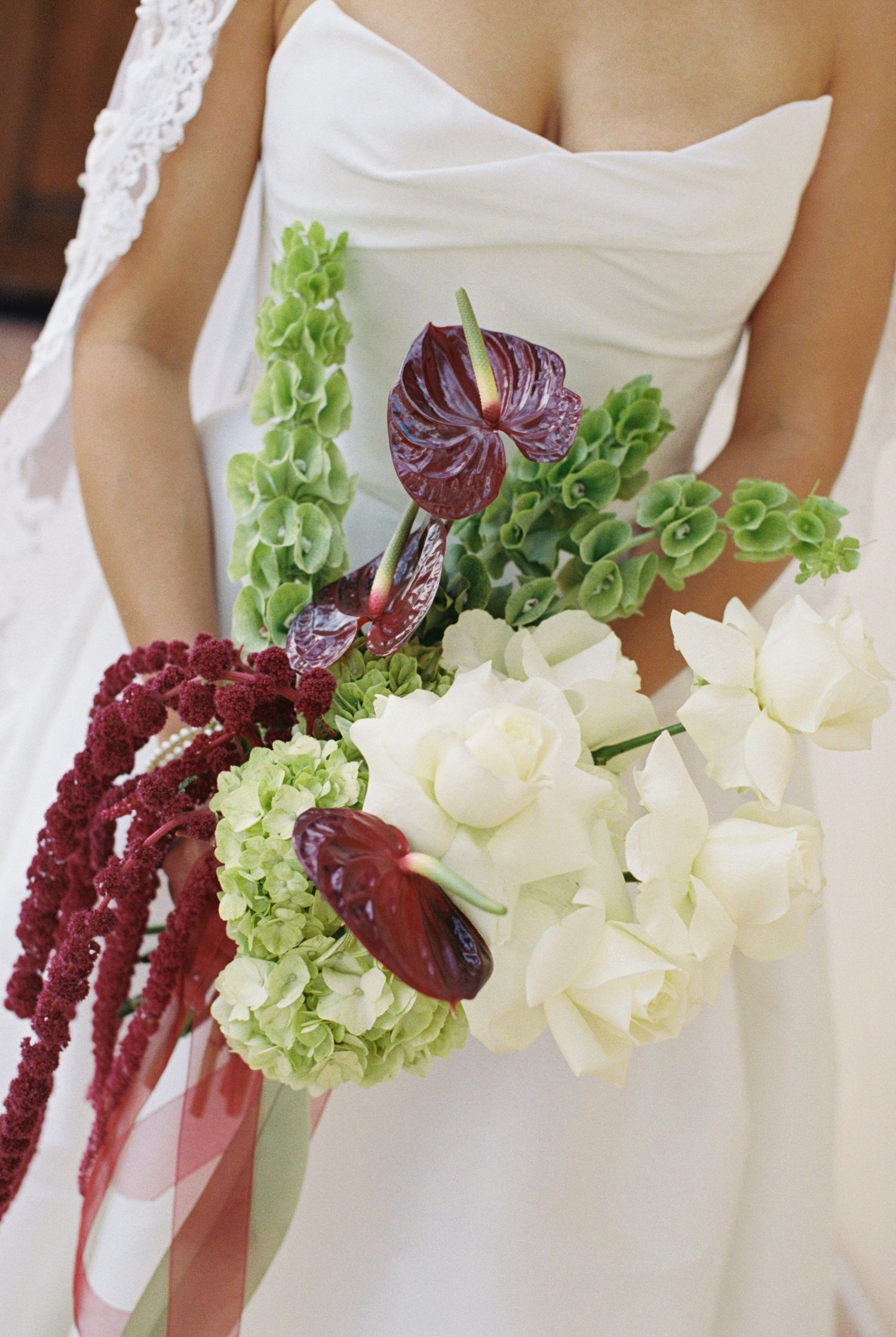 Bride holding elegant wedding bouquet with white tulips, burgundy anthuriums, green hydrangeas, and cascading amaranthus.