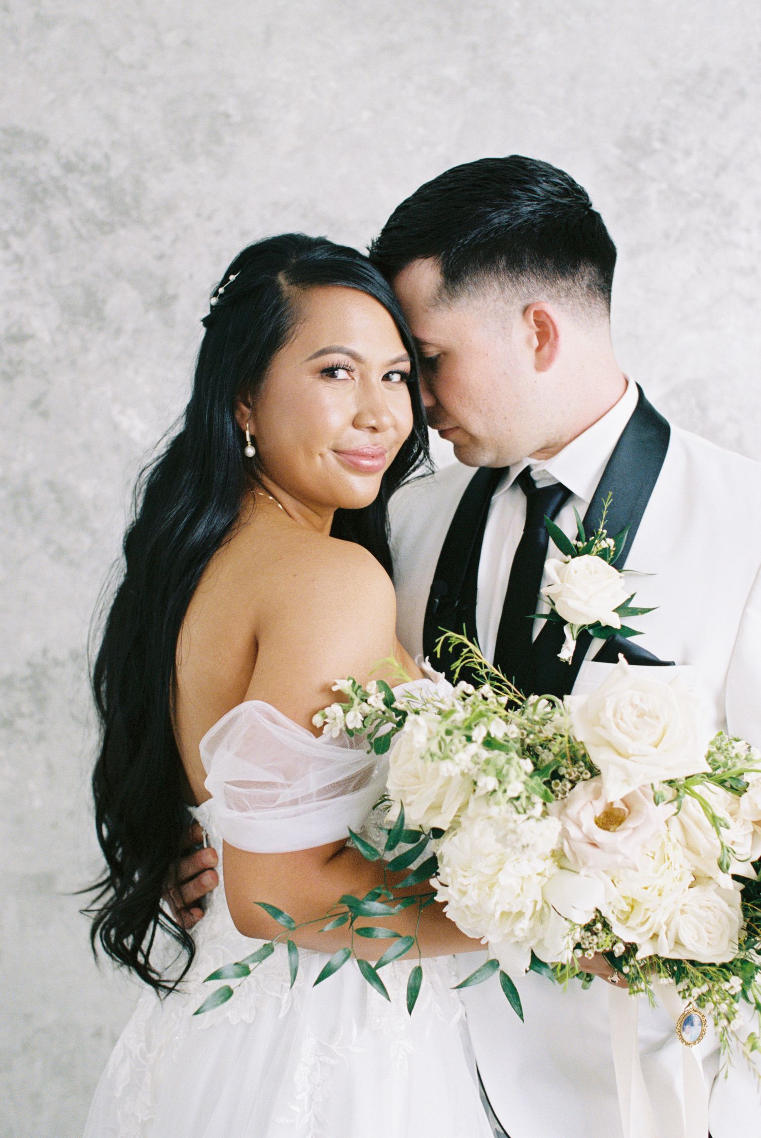 Bride in off-shoulder gown holds white floral bouquet while groom in tuxedo embraces her against gray backdrop.
