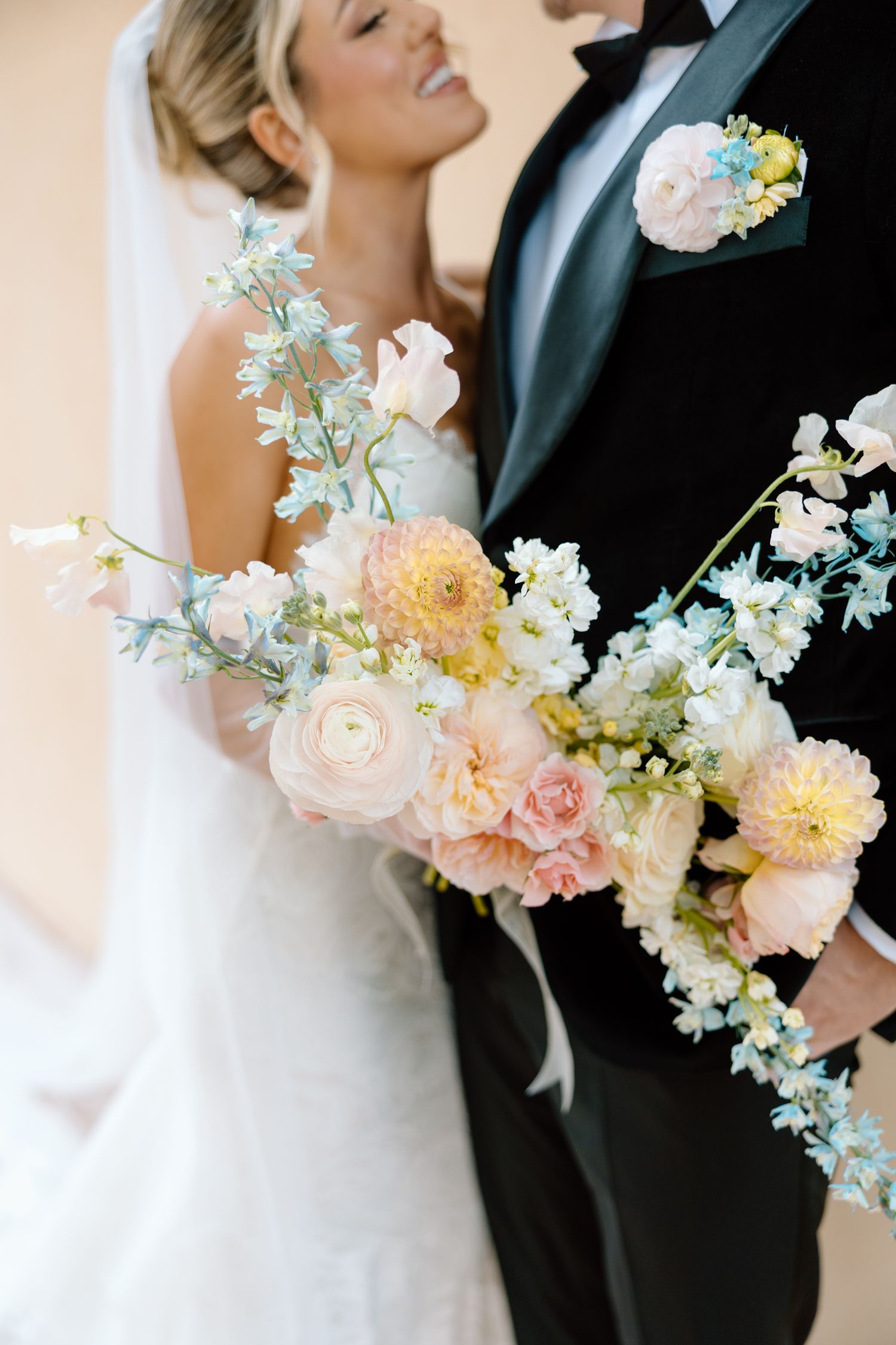 A romantic wedding couple embraces while holding a pastel bouquet of pink and white flowers against a light background.