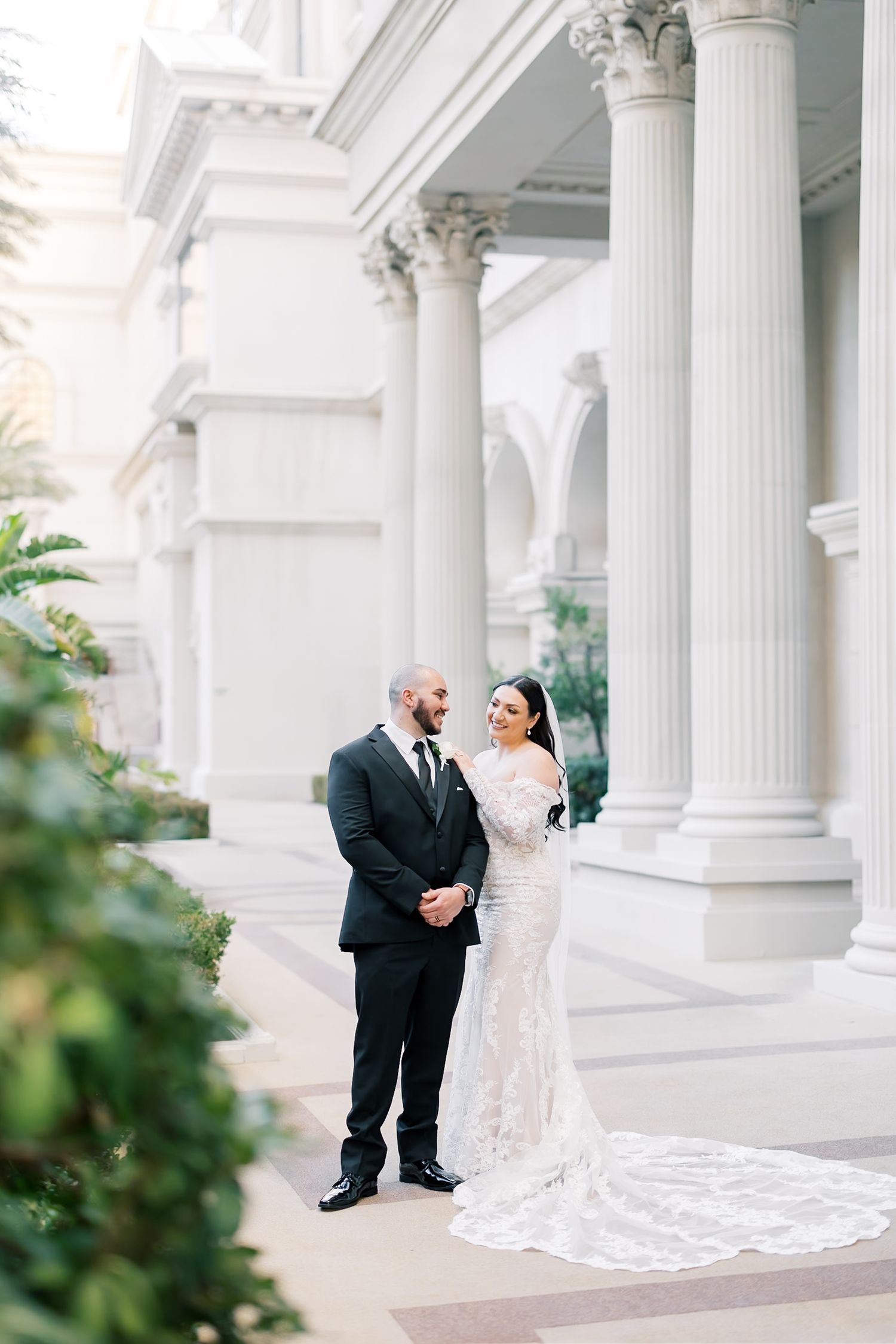 Bride in lace gown and groom in black tuxedo pose before grand white columns at elegant wedding venue.