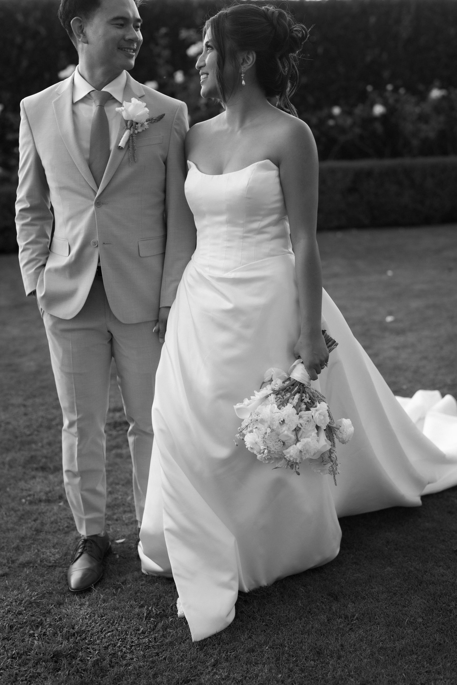 Black and white photo of a smiling bride holding a bouquet and groom in a suit walking together on grass.