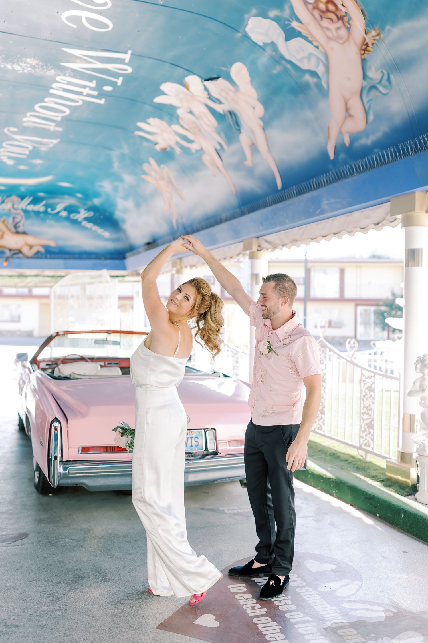 Bride and groom dancing in front of a pink convertible at a Las Vegas wedding chapel with cherub murals.