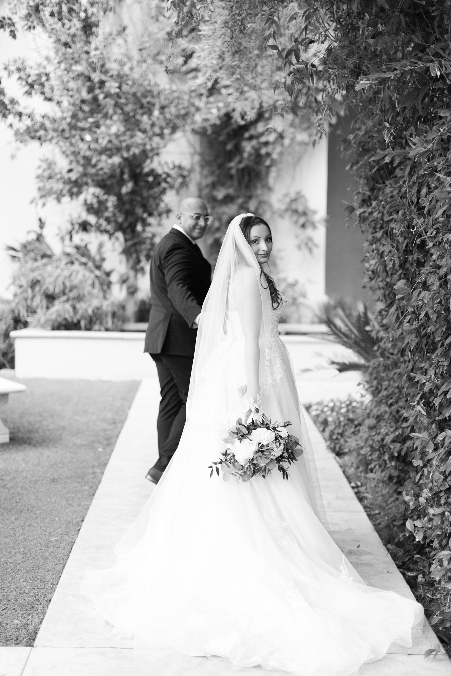 Black and white photo of a bride in a flowing gown with veil and bouquet, standing with a groom in a garden.