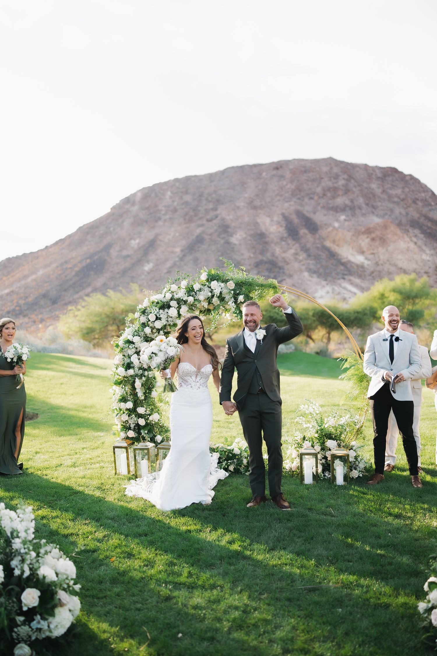 Joyful newlyweds celebrate under a floral arch on a green lawn with a stunning desert mountain backdrop.
