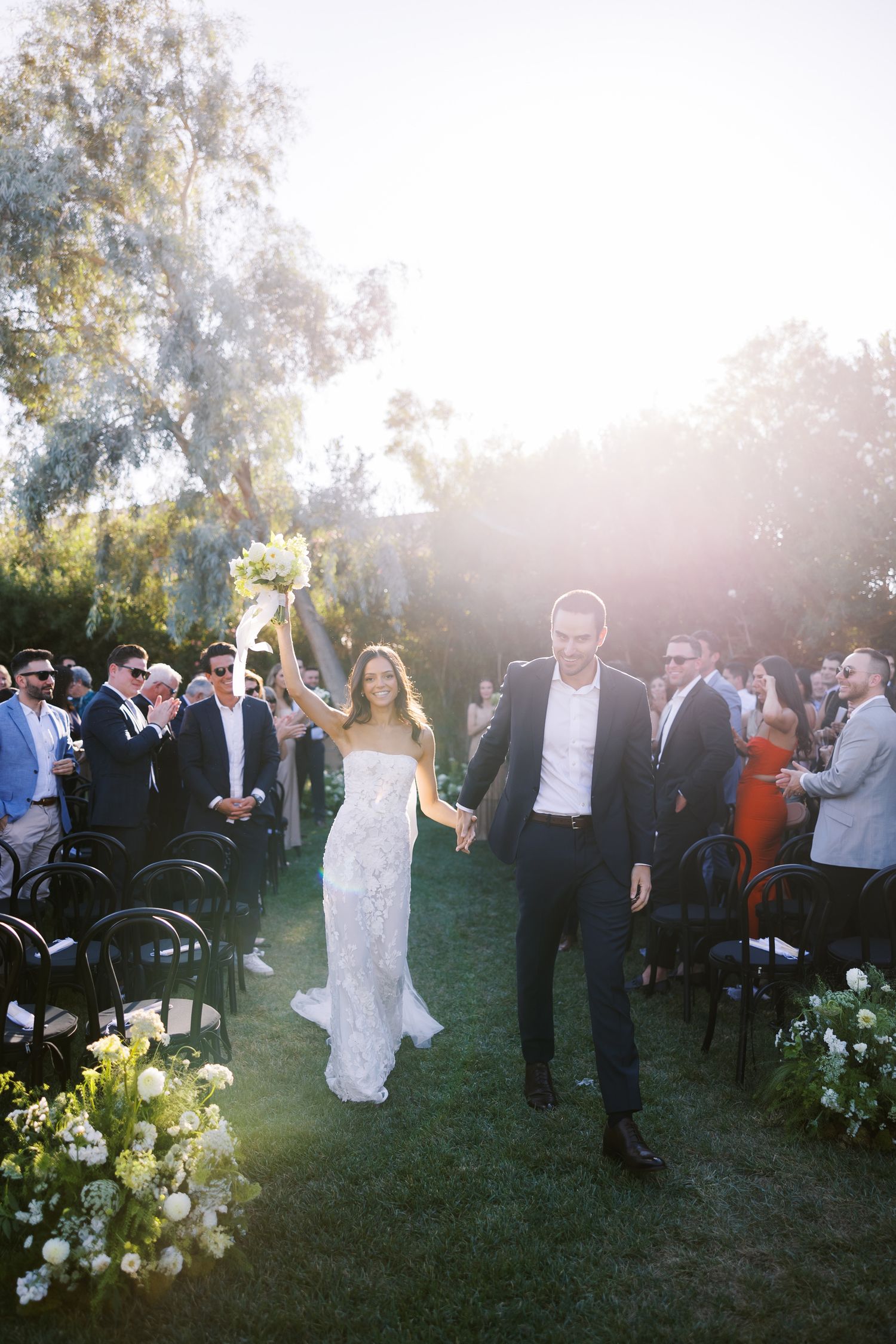 Bride and groom walk down outdoor aisle after ceremony, sun-kissed garden setting with floral arrangements and guests.
