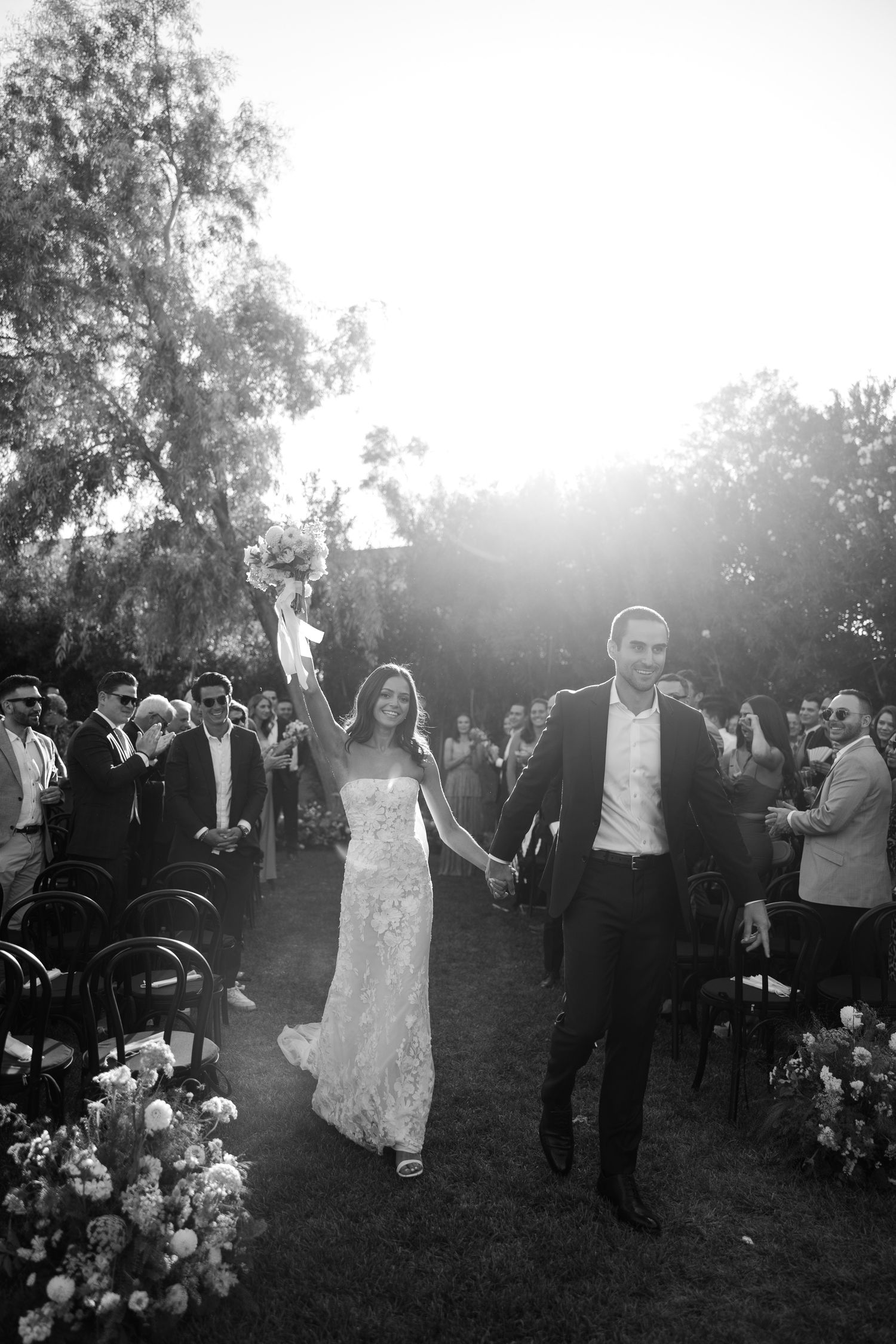 A wedding party walks down an outdoor aisle at sunset with guests looking on from both sides.