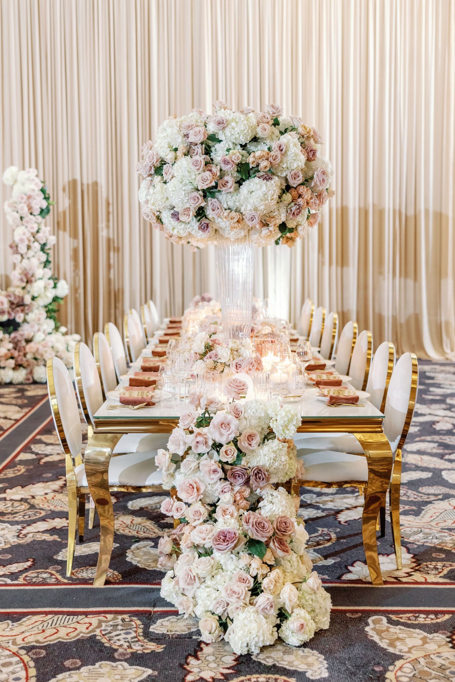 Romantic wedding reception table with cascading blush and white rose runner, tall floral centerpiece, and gold chairs.