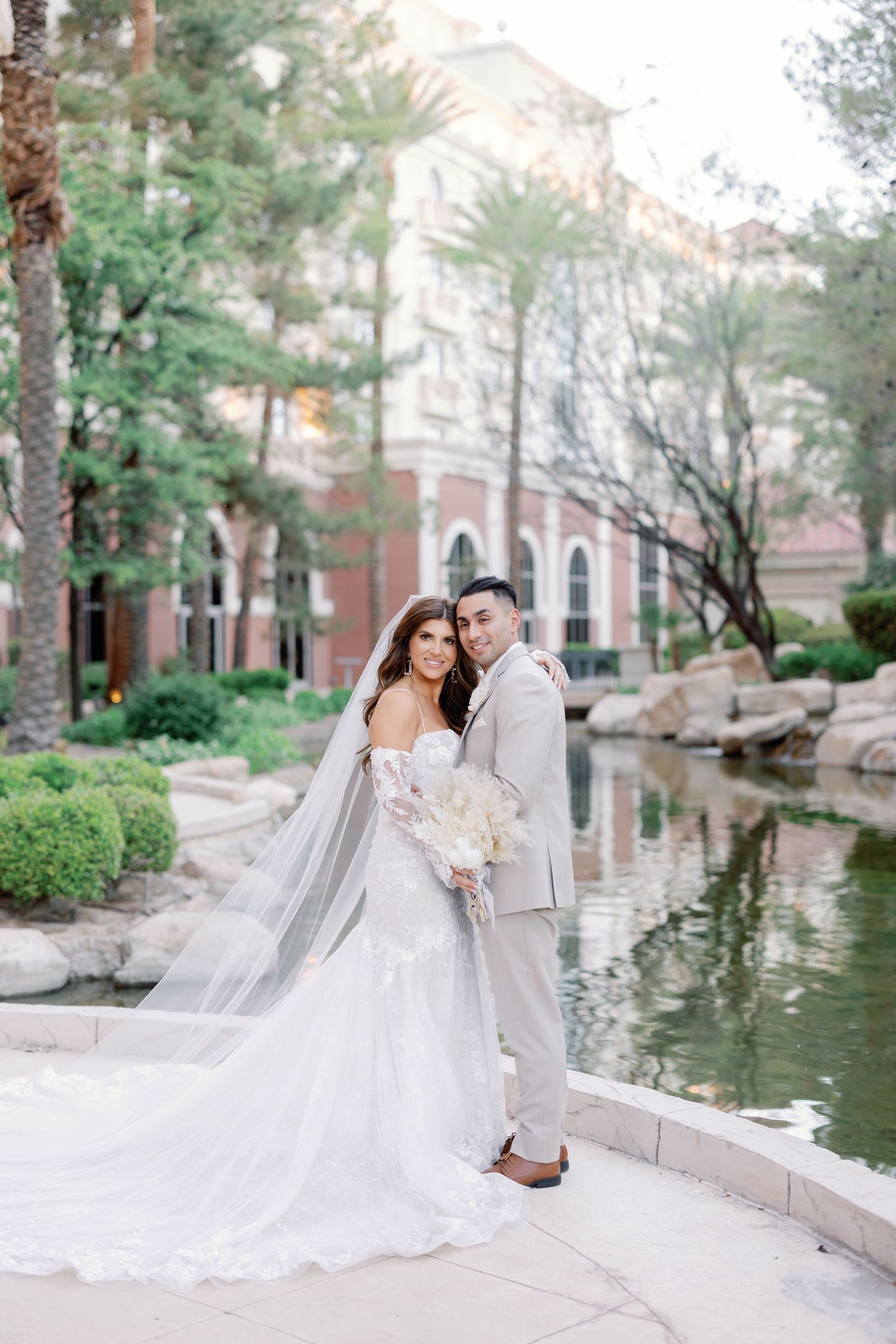 Bride in a flowing white gown with long veil and groom in gray suit posing by a serene garden pond.