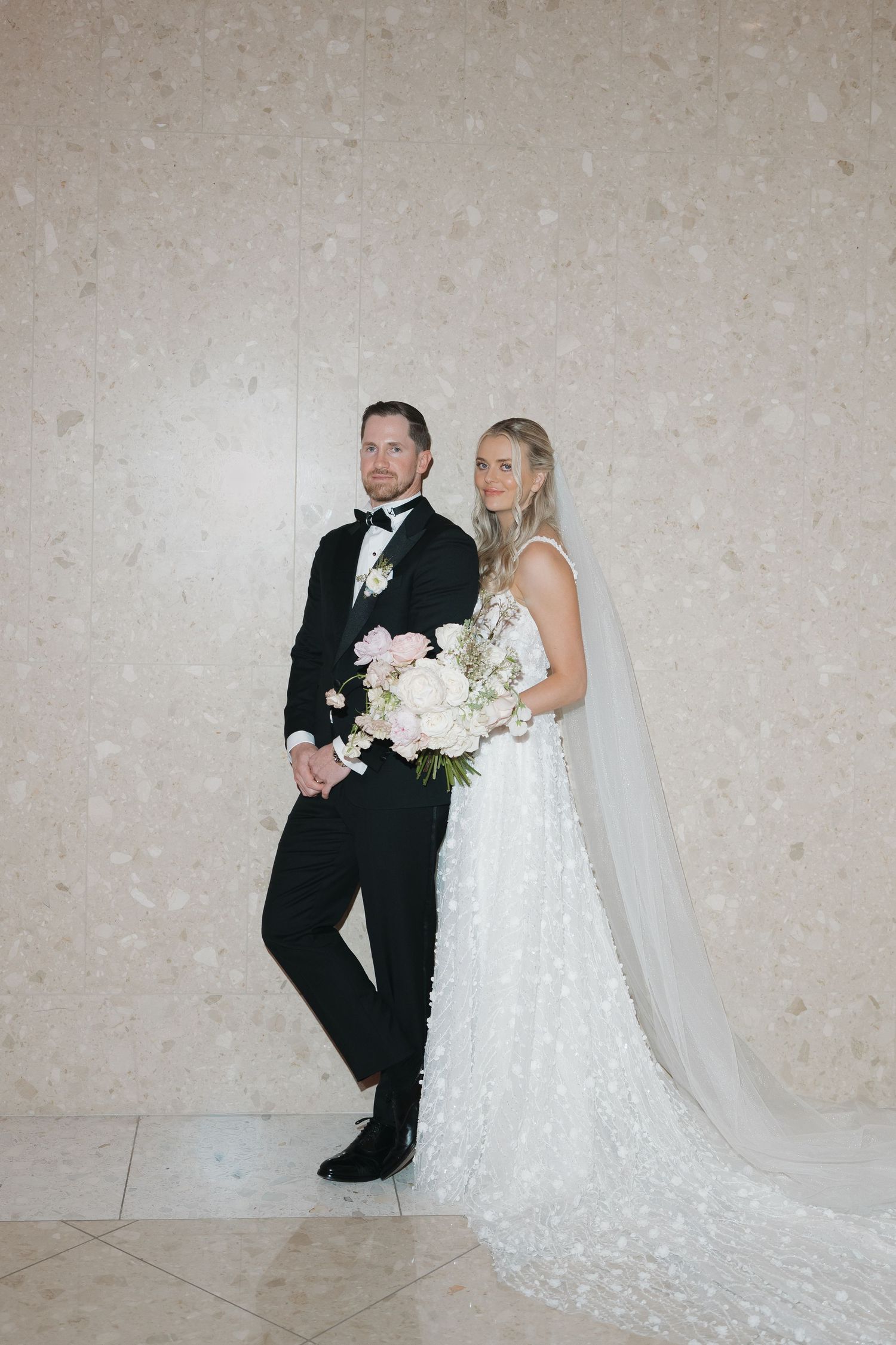 Elegant bride and groom portrait against marble wall, bride holding white and pink bouquet.