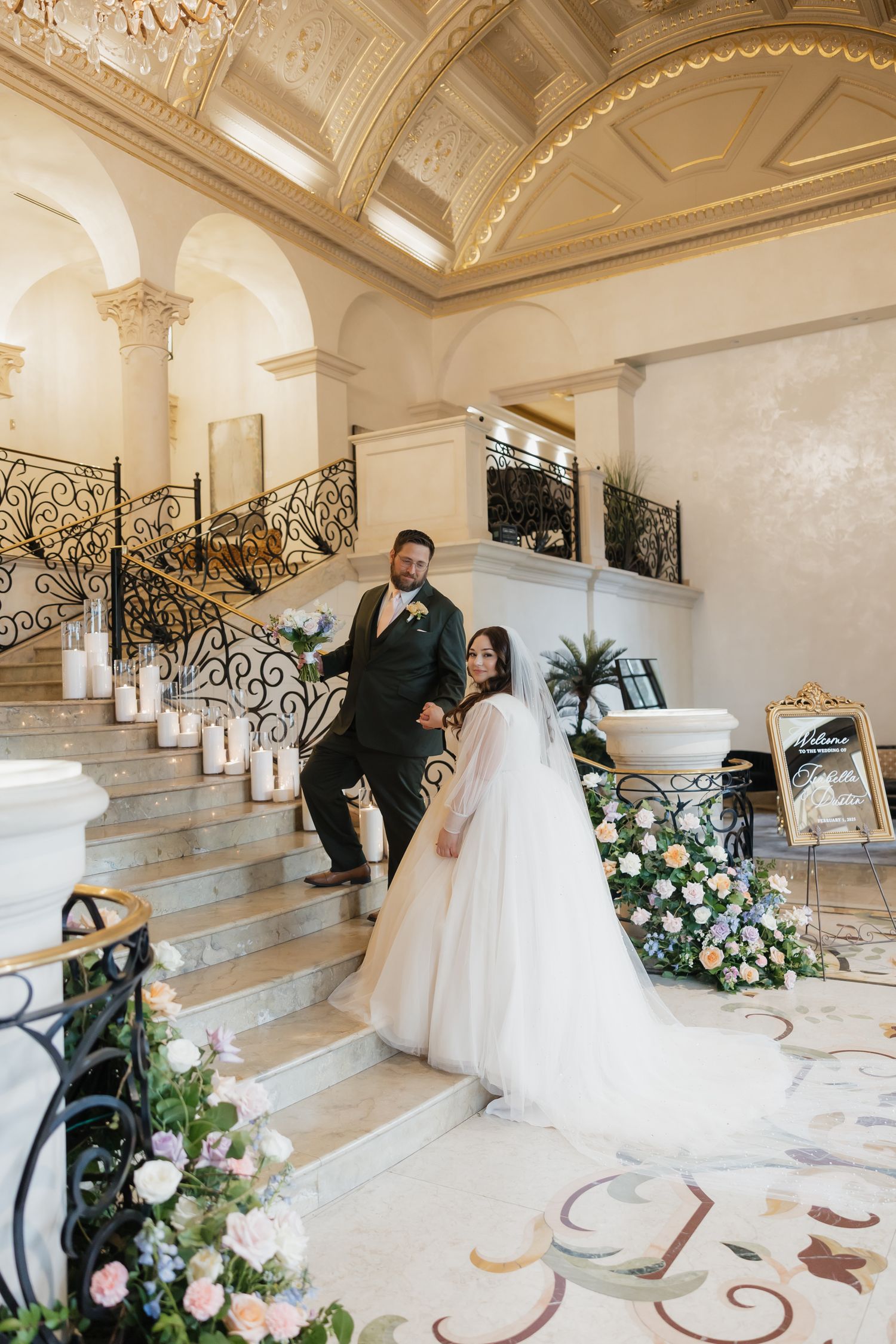 Bride and groom pose on grand staircase with floral arrangements in elegant venue.