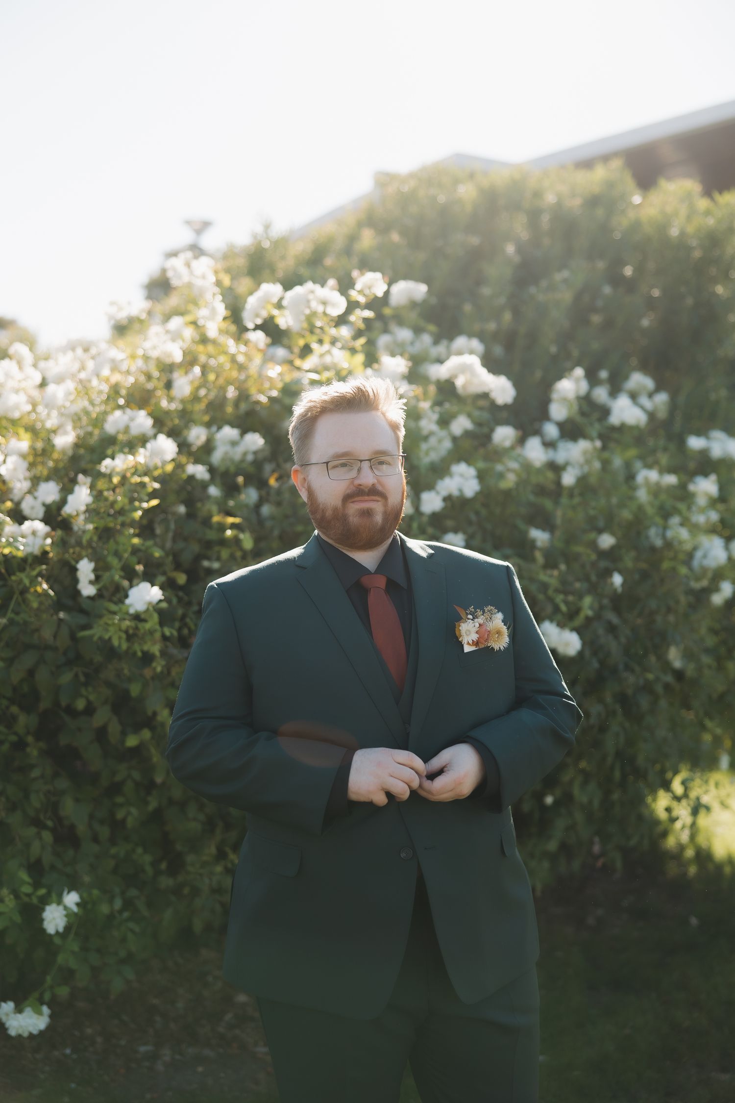 Groom in dark green suit with burgundy tie and floral boutonniere standing before white rose bushes.