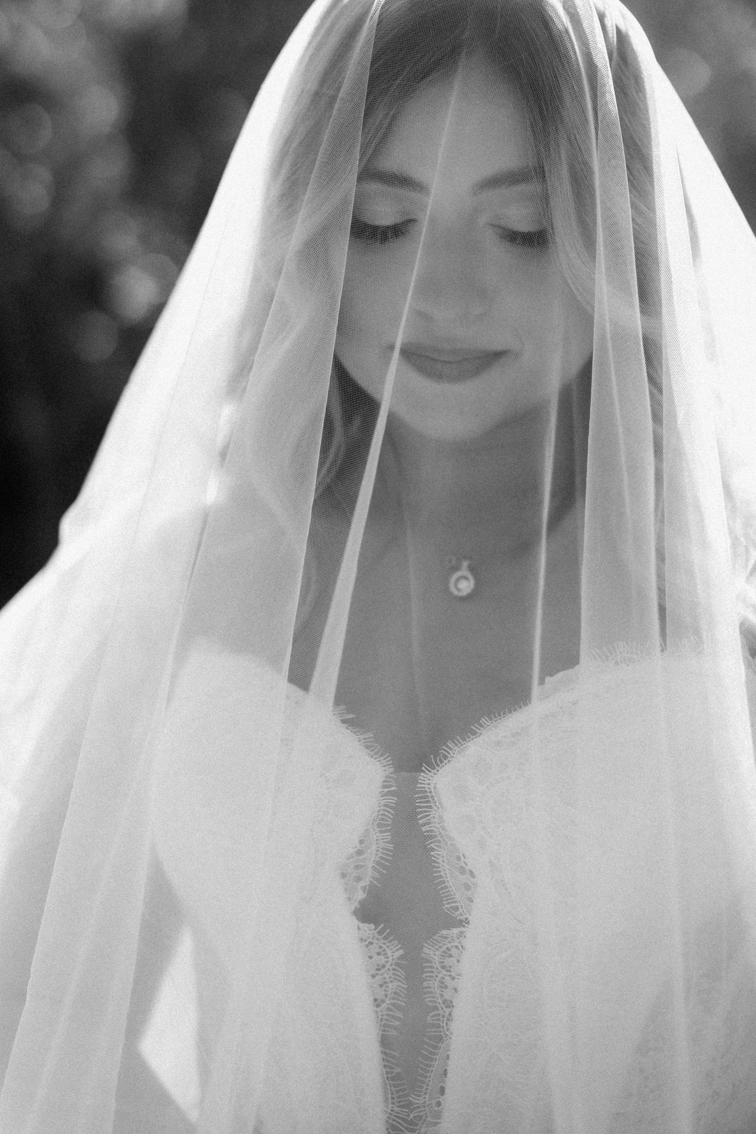 Black and white portrait of a bride wearing a veil draped over her face, lace wedding dress, and delicate necklace.