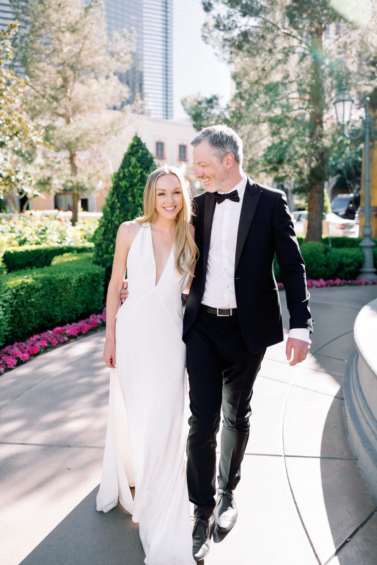 Bride in white gown and groom in black tuxedo walking together outdoors at an elegant garden venue.