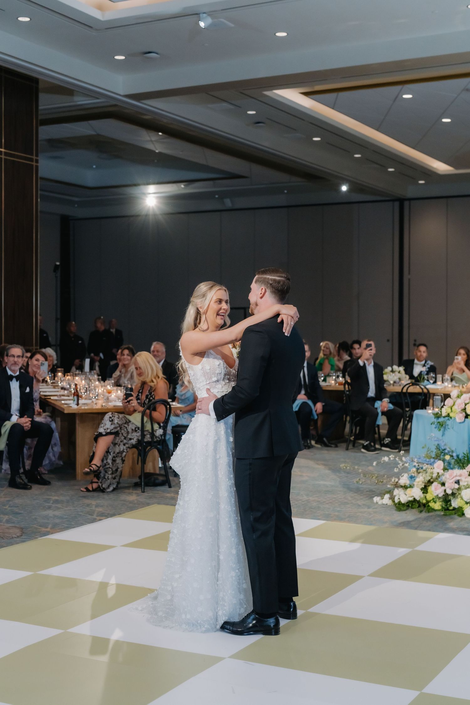 Bride and groom share their first dance on a checkered dance floor surrounded by seated wedding guests.
