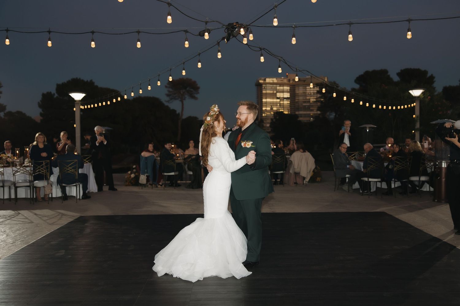 Couple shares first dance on outdoor floor under string lights at evening wedding reception.