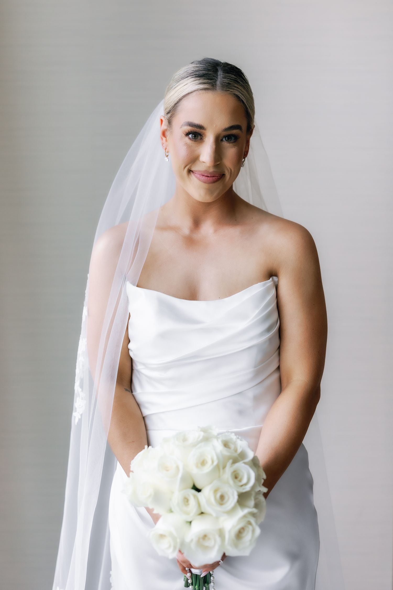 Elegant bride in strapless white gown and veil holding white rose bouquet, smiling softly against neutral background.