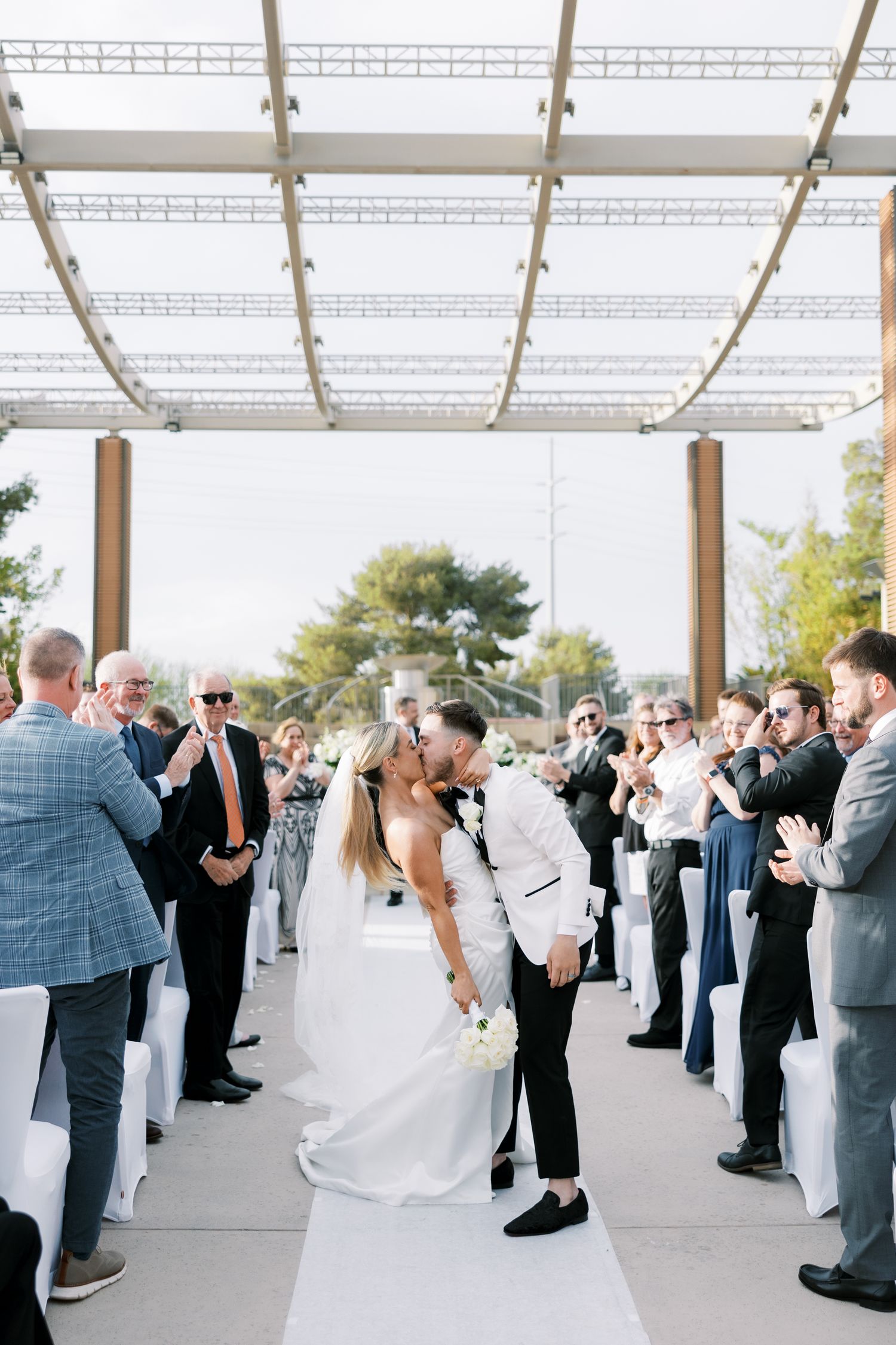 Bride and groom share a kiss at the end of the aisle during outdoor wedding ceremony with guests applauding.