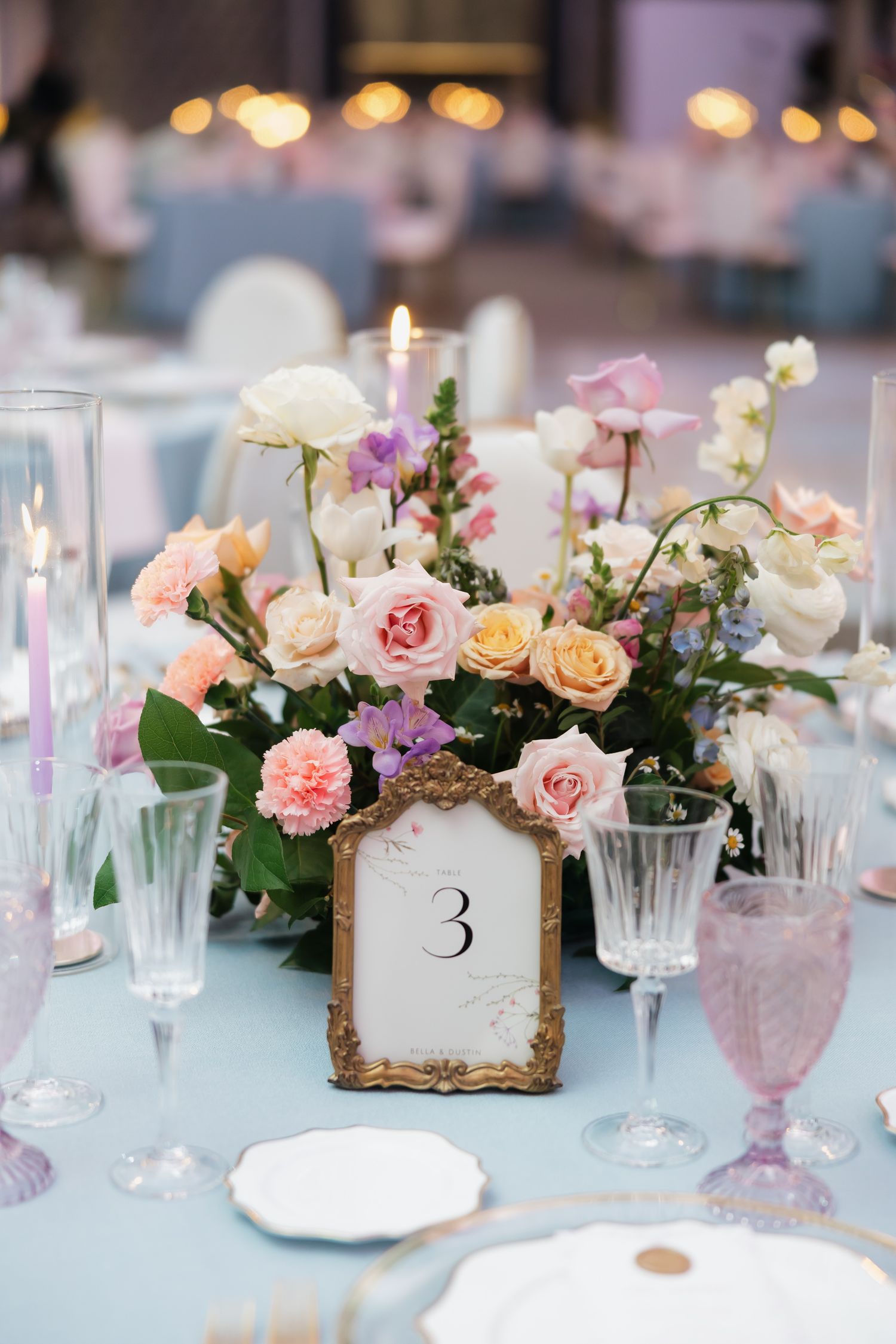 Elegant wedding table centerpiece with pastel roses, candles, crystal glasses, and a gold-framed table number 3 on a blue tablecloth.