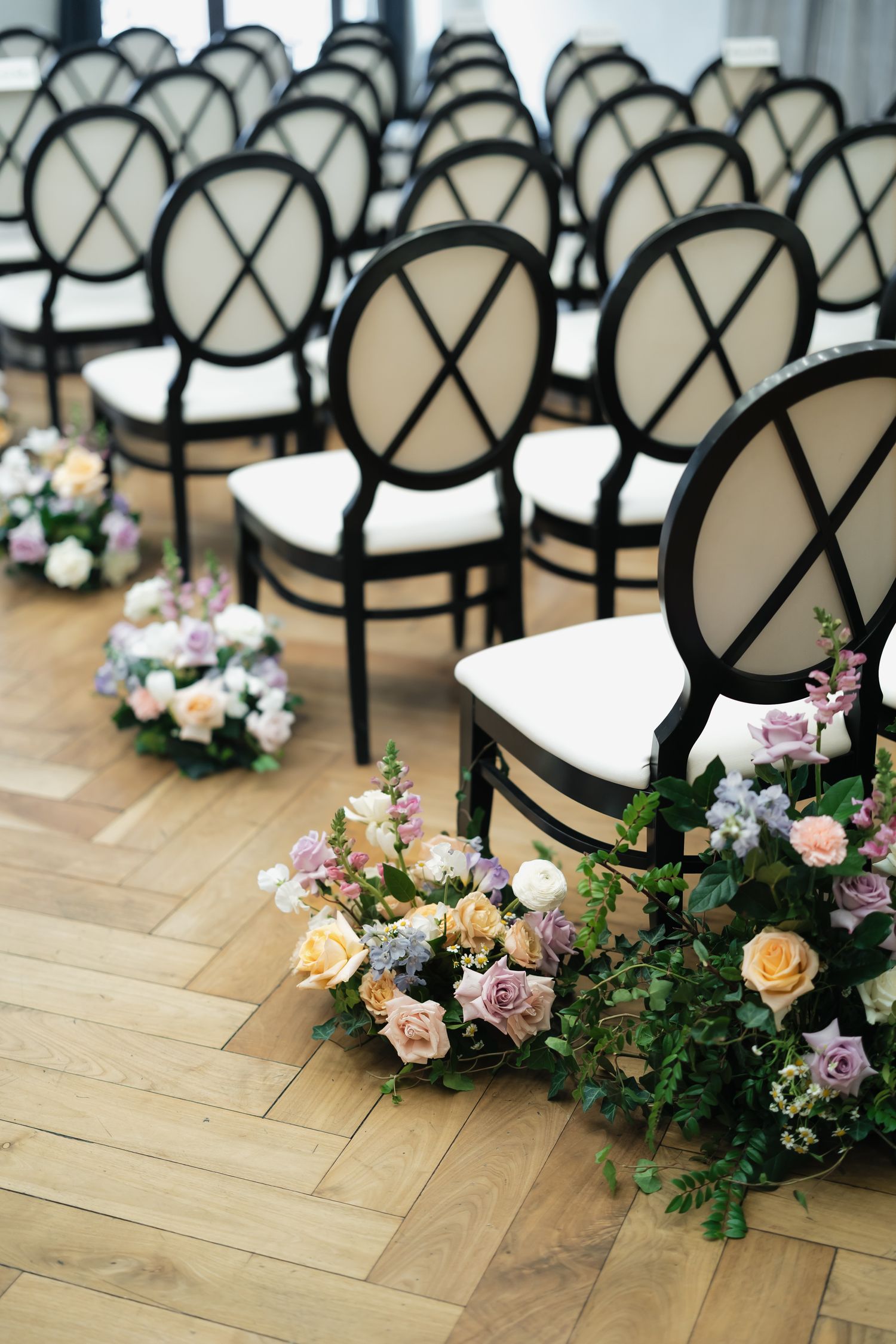 Elegant wedding ceremony venue with black and white chairs arranged in rows on herringbone wood floors, adorned with floral arrangements.