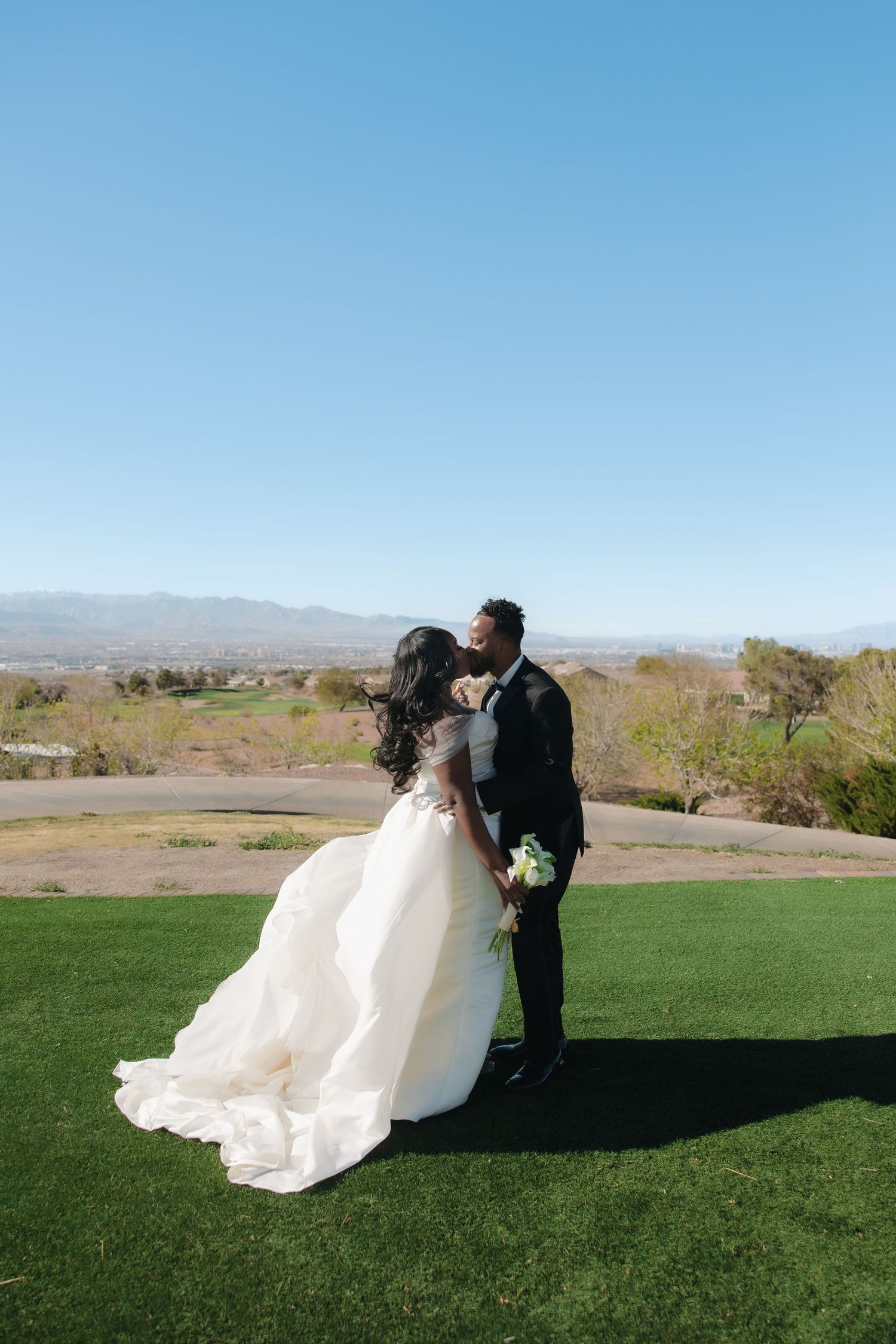 Bride and groom share a romantic kiss on green lawn with scenic mountain valley backdrop on sunny wedding day.