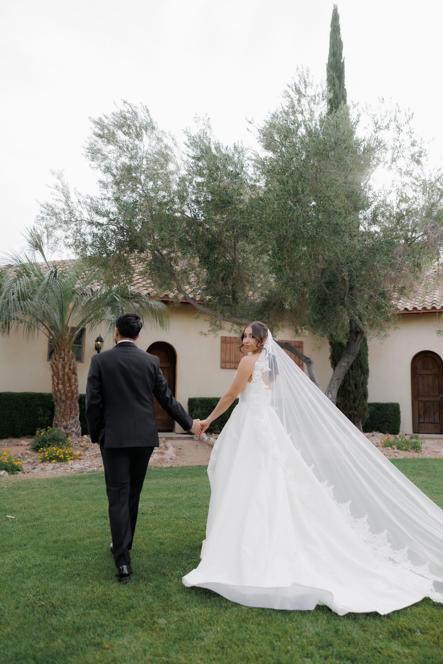 Bride and groom walk hand-in-hand across green lawn, bride's cathedral veil trailing behind, near a Spanish-style villa.