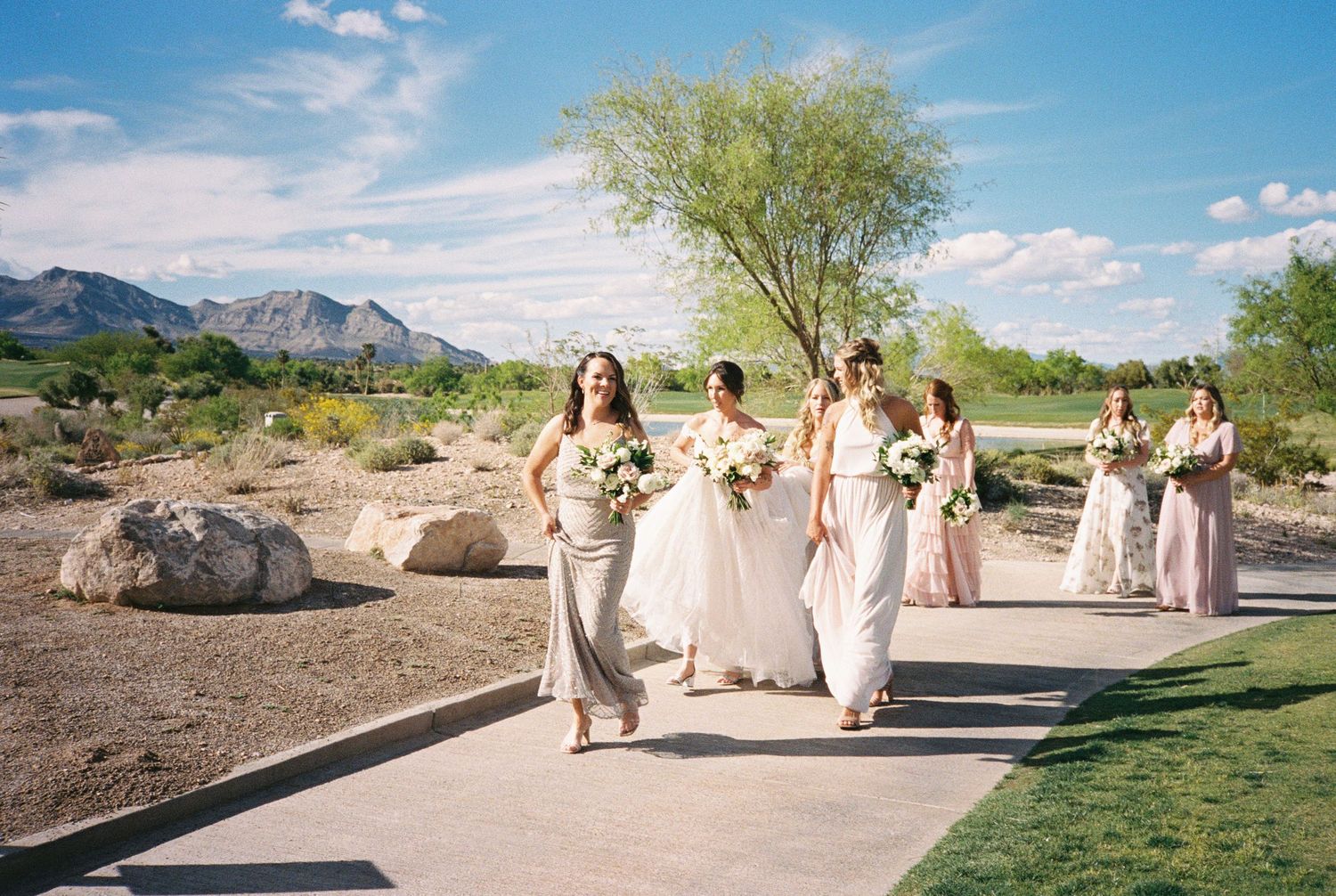 Bridesmaids in champagne and blush gowns walk along a path at an outdoor desert wedding venue with mountains.