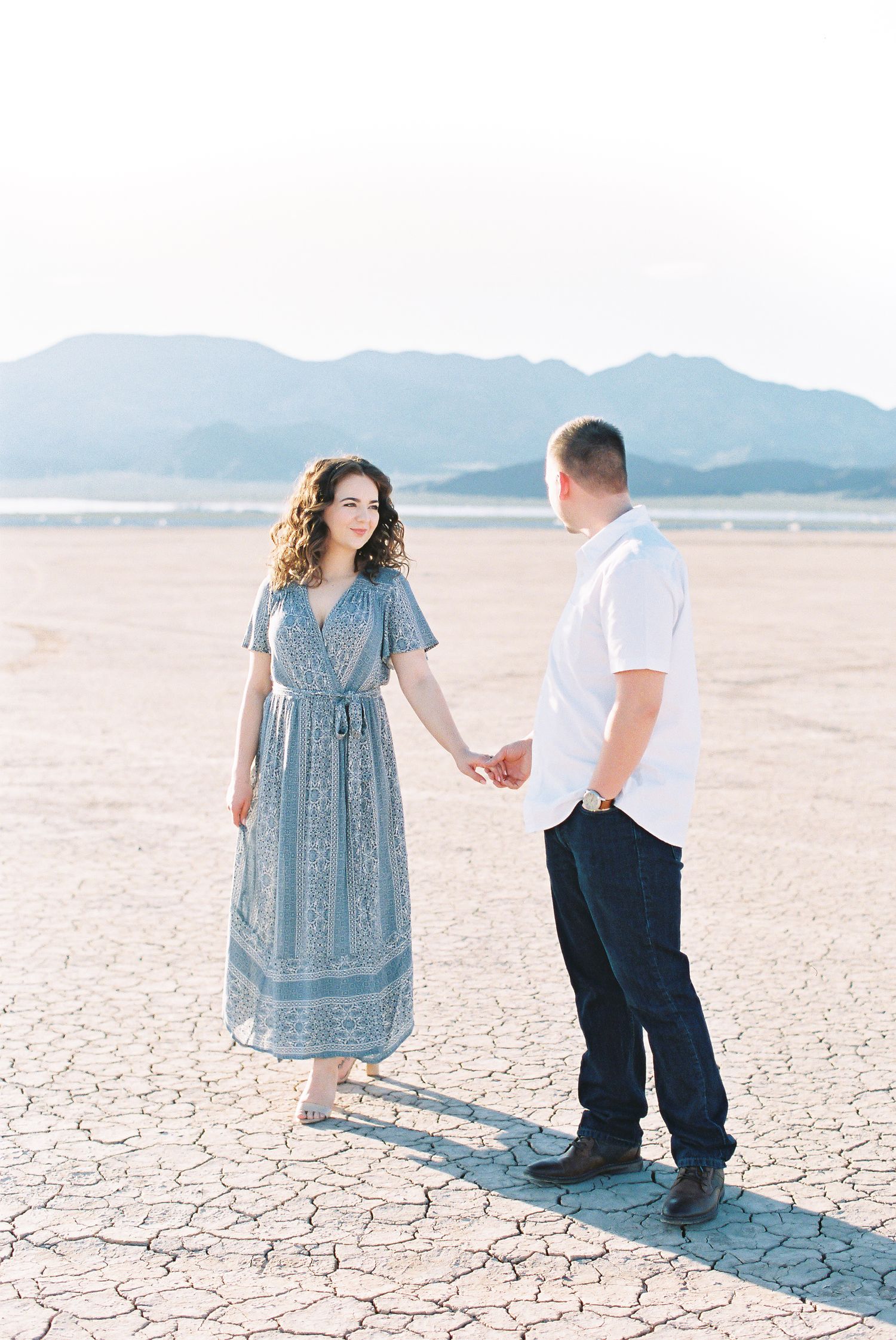 Couple holding hands in a vast dry lakebed with mountains, woman in blue floral dress, man in white shirt.