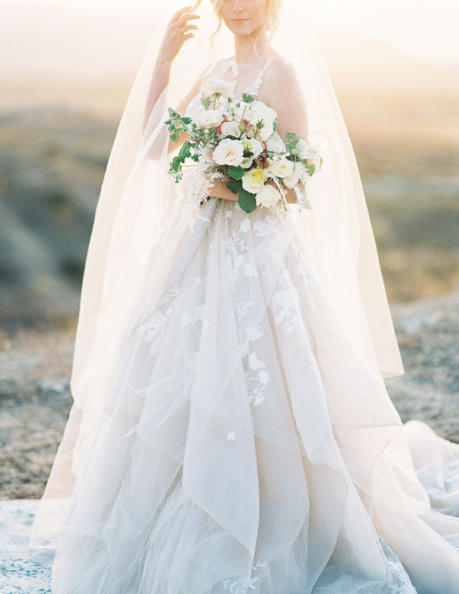 Bride in a flowing white ball gown with floral appliques holds a white bouquet, wearing a veil in a sunlit outdoor setting.